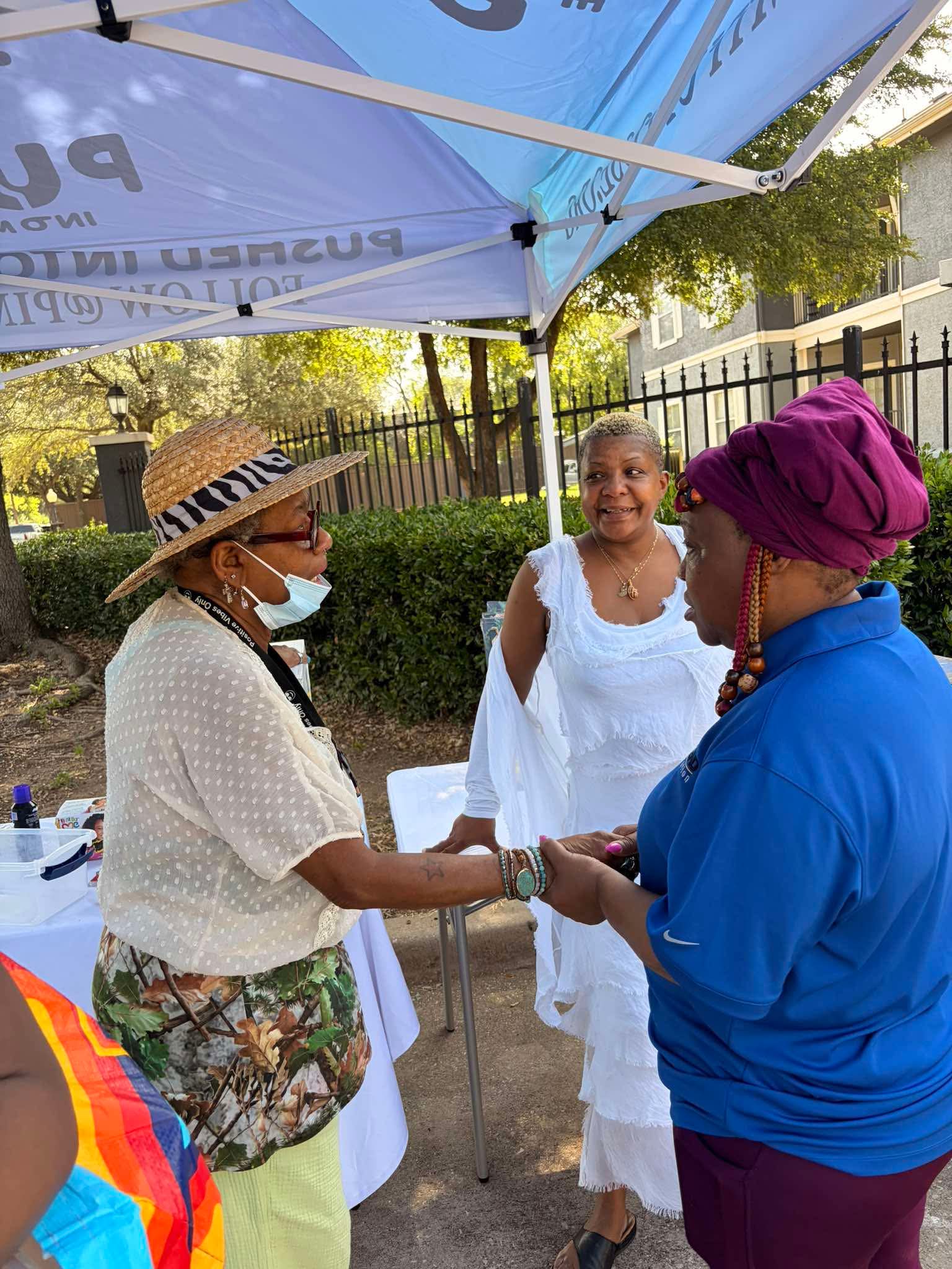Three women outdoors under a blue canopy, two women shaking hands, one woman smiling, trees and a fence in background.