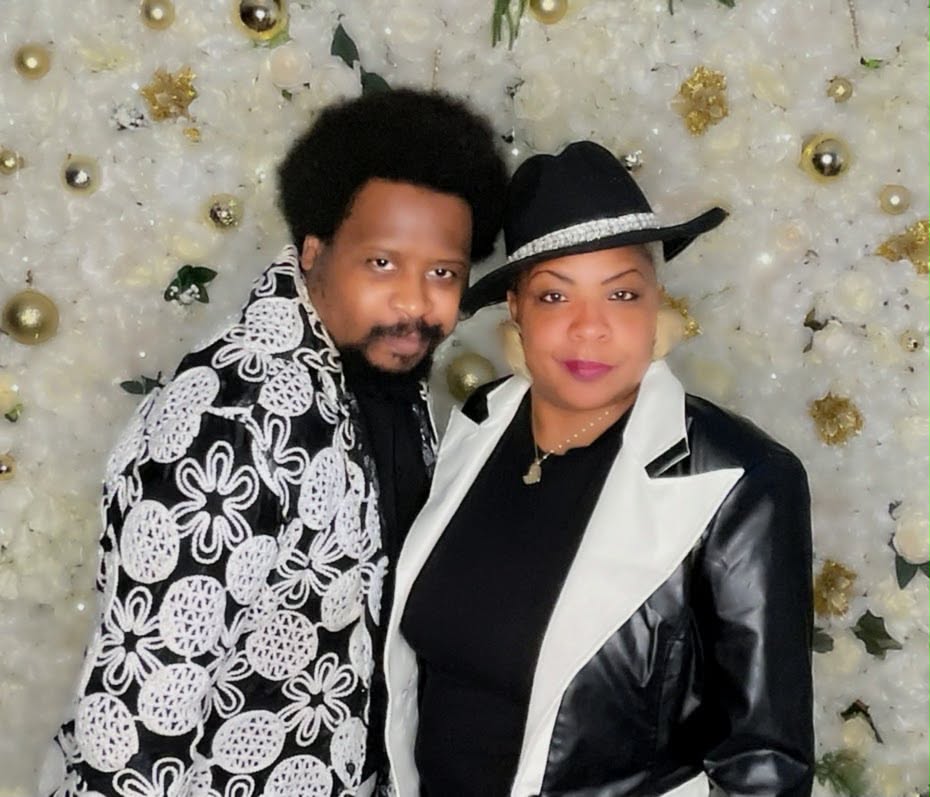 Two people standing close together in front of a decorated white background with gold accents.