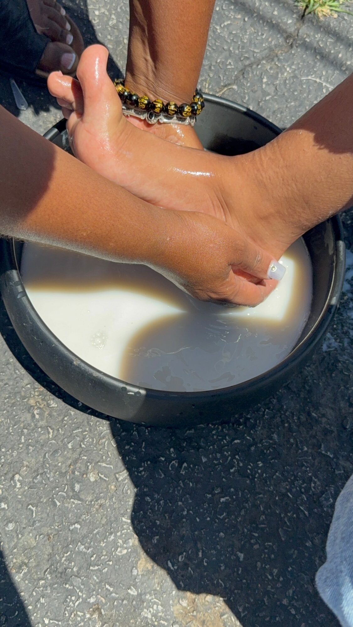 Person's foot being washed in a black basin with soapy water, outdoors on pavement, with a bracelet visible on wrist.