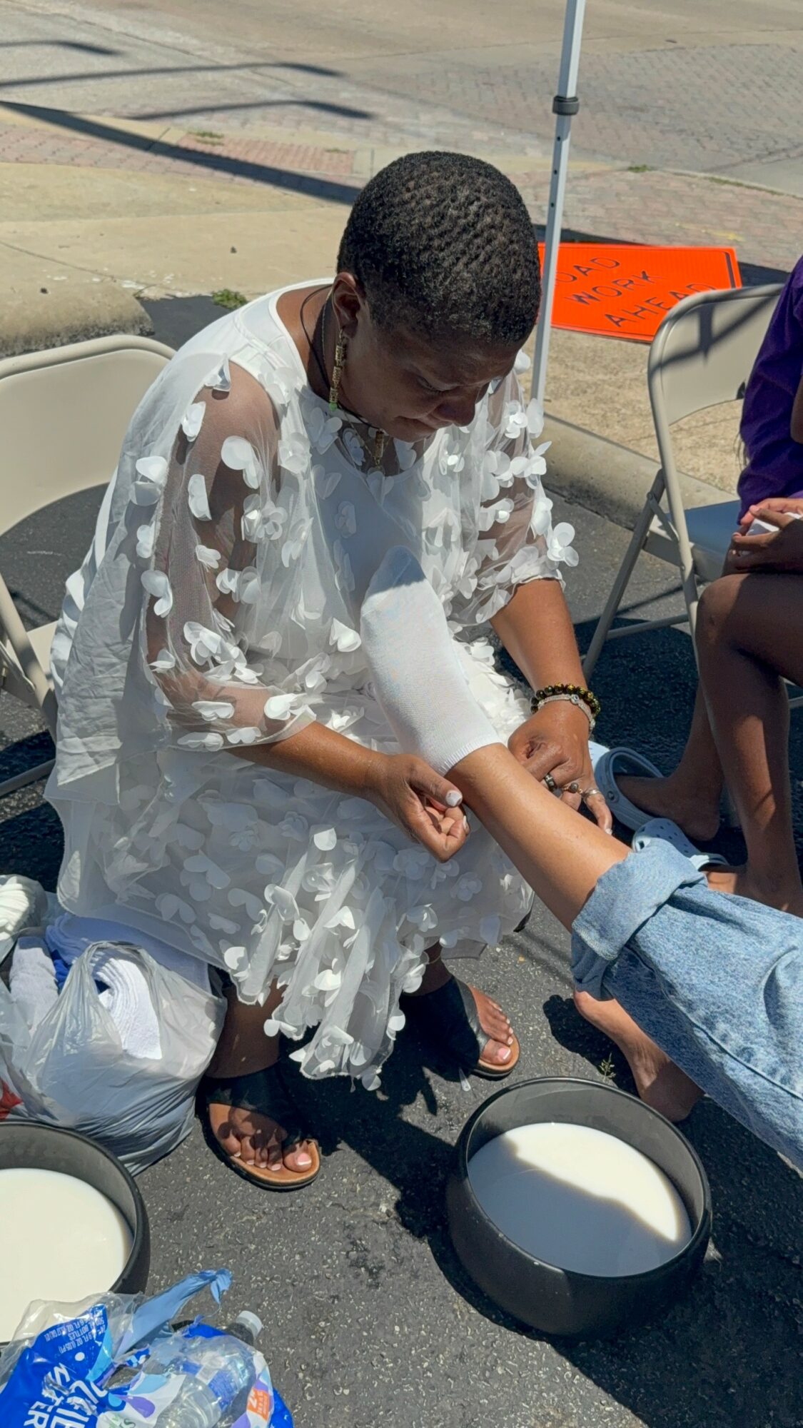 Person in a white dress with foam on arms, sitting outdoors, holding a person's foot over a bowl, others nearby.