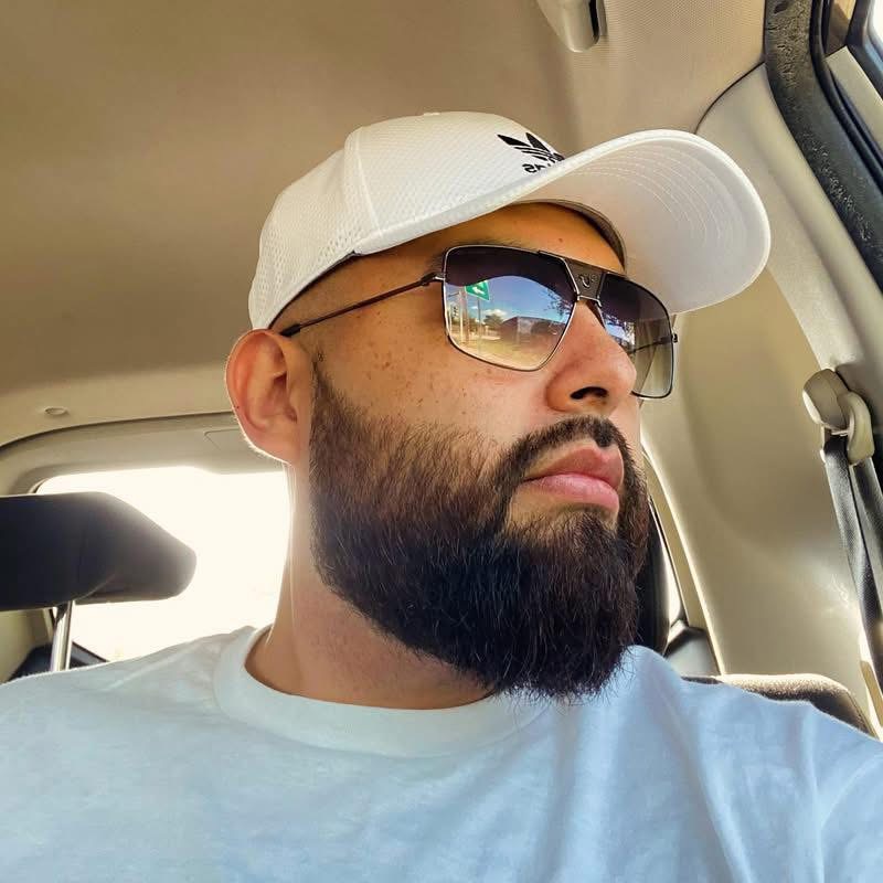 Man with beard wearing sunglasses and a white cap, sitting in a vehicle, looking to the side.