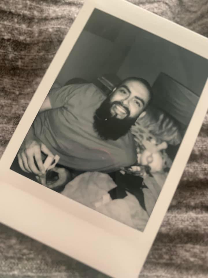 Smiling man with a beard lying on a bed, holding a phone, in a black and white photo.