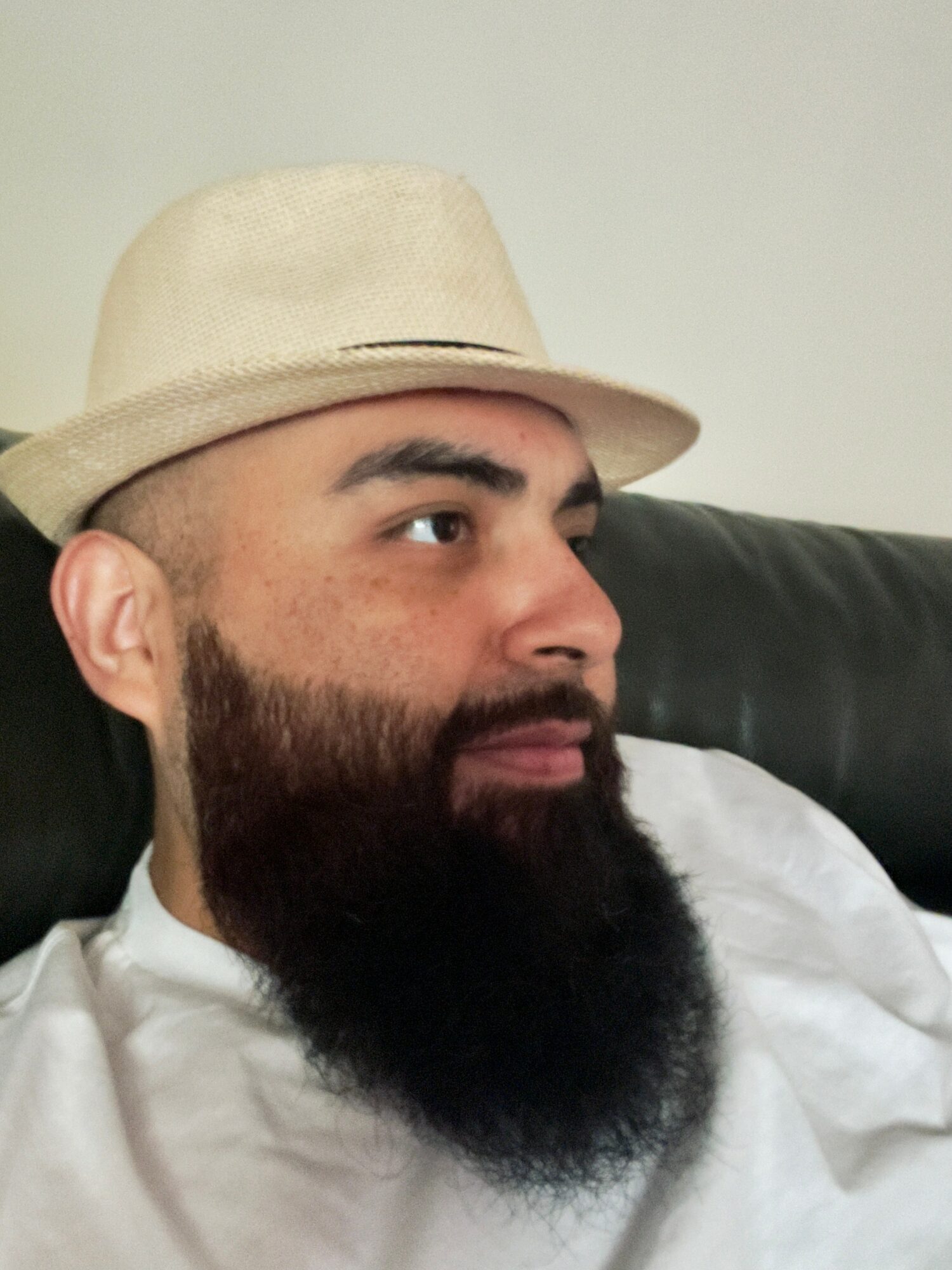 Man with a beard wearing a beige hat and white shirt, sitting on a black couch, looking to the right.