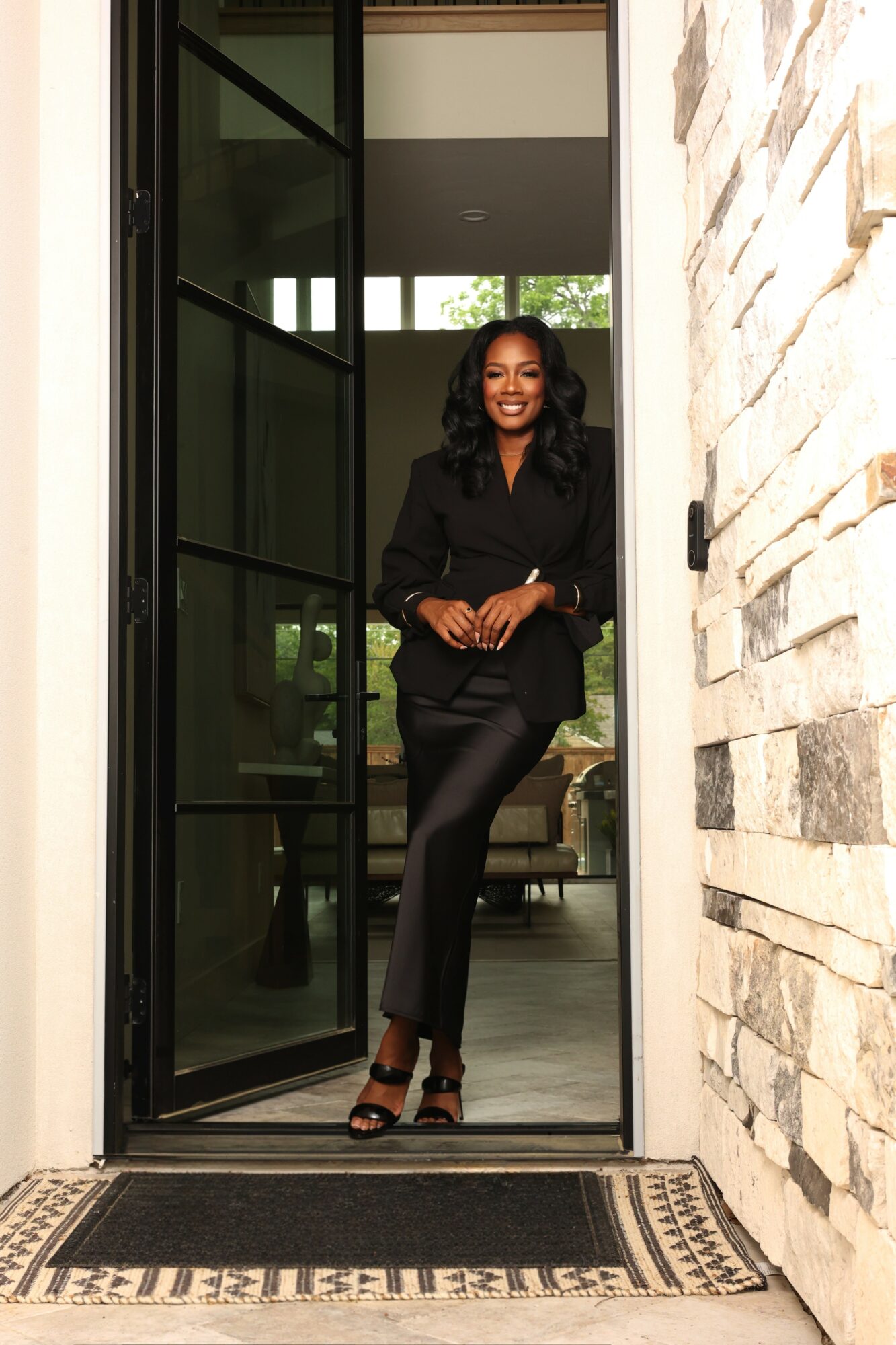 Woman standing in doorway, smiling, wearing black outfit, with a stone wall on the right and glass door behind.