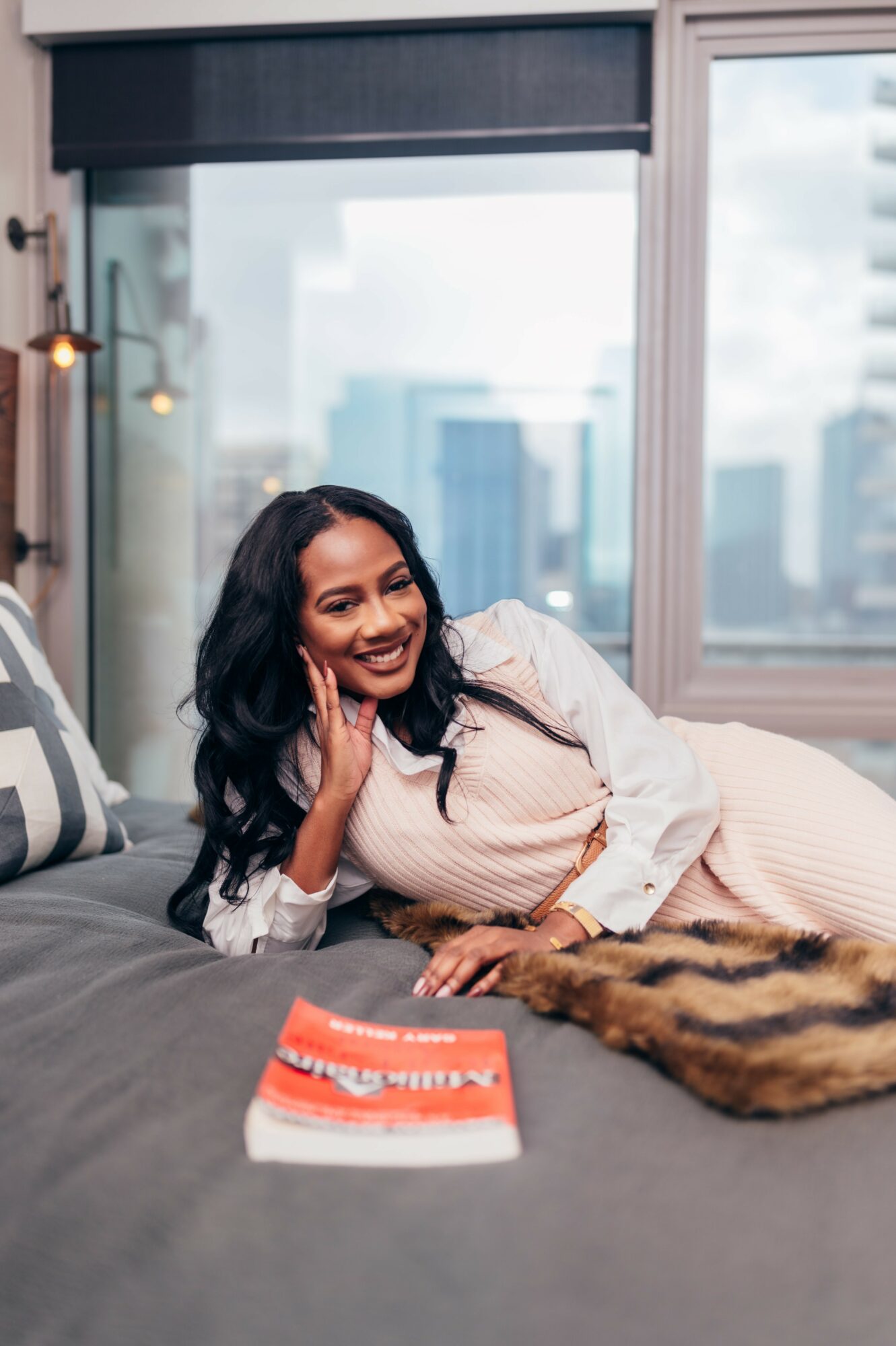 Woman with dark hair smiling and lying on a bed in a modern apartment, city skyline visible through large window.