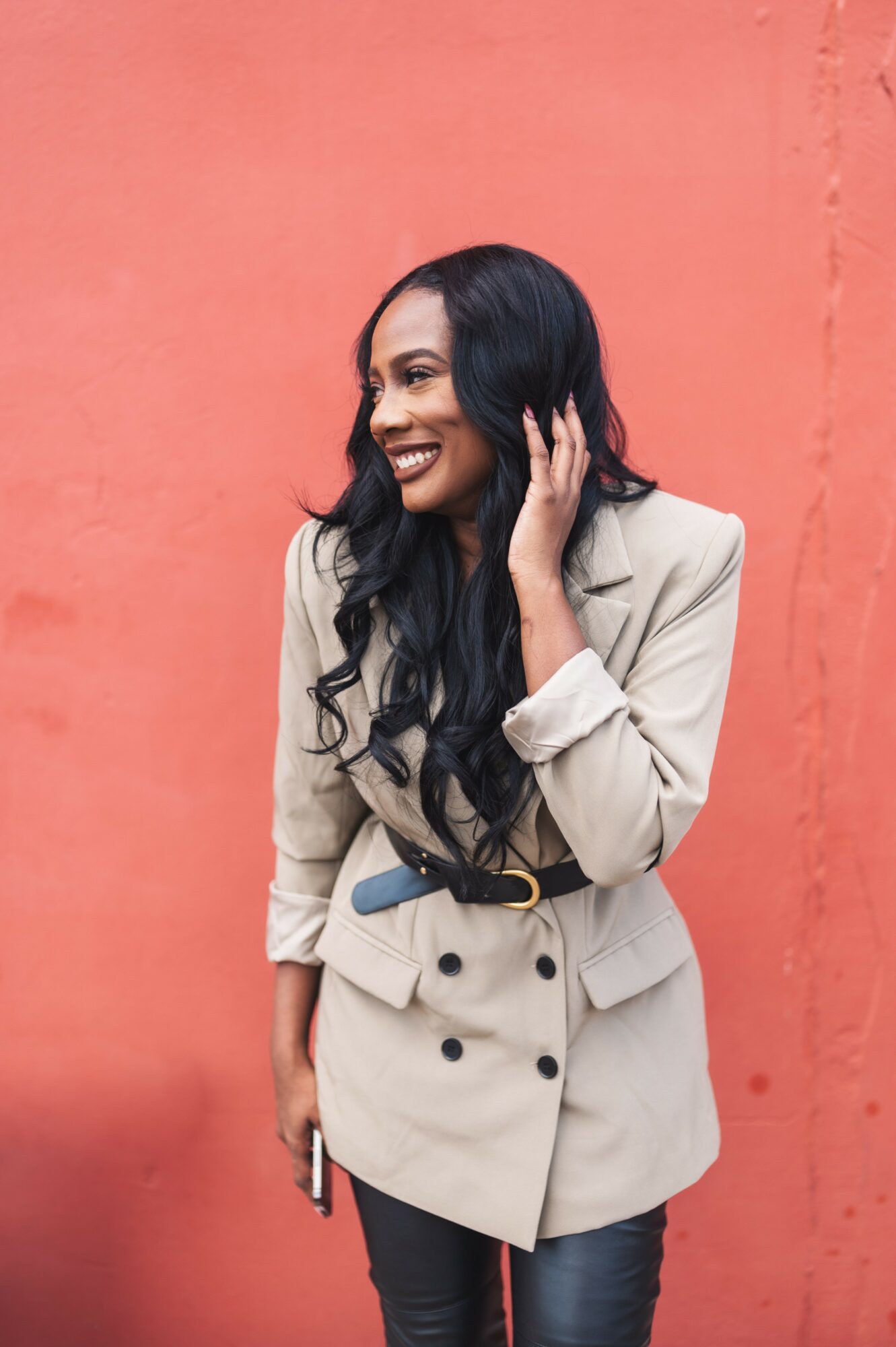 Woman with long black hair smiling, wearing a beige blazer and dark pants, standing against a pink wall.