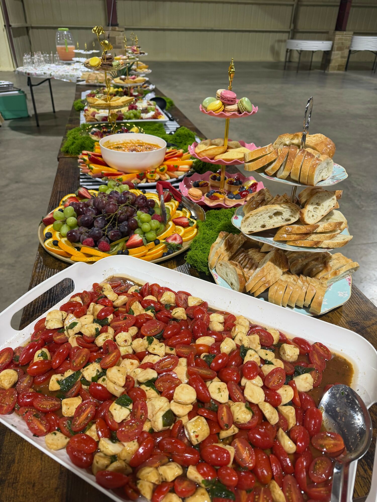 Long table with various dishes including pasta, bread, fruit, and desserts, set in a spacious indoor area.