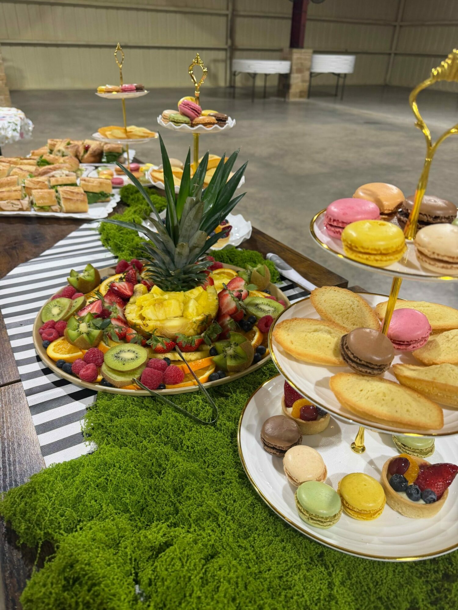 Assorted desserts and fruits displayed on a table with green moss, including macarons, cake slices, and a pineapple centerpiece.