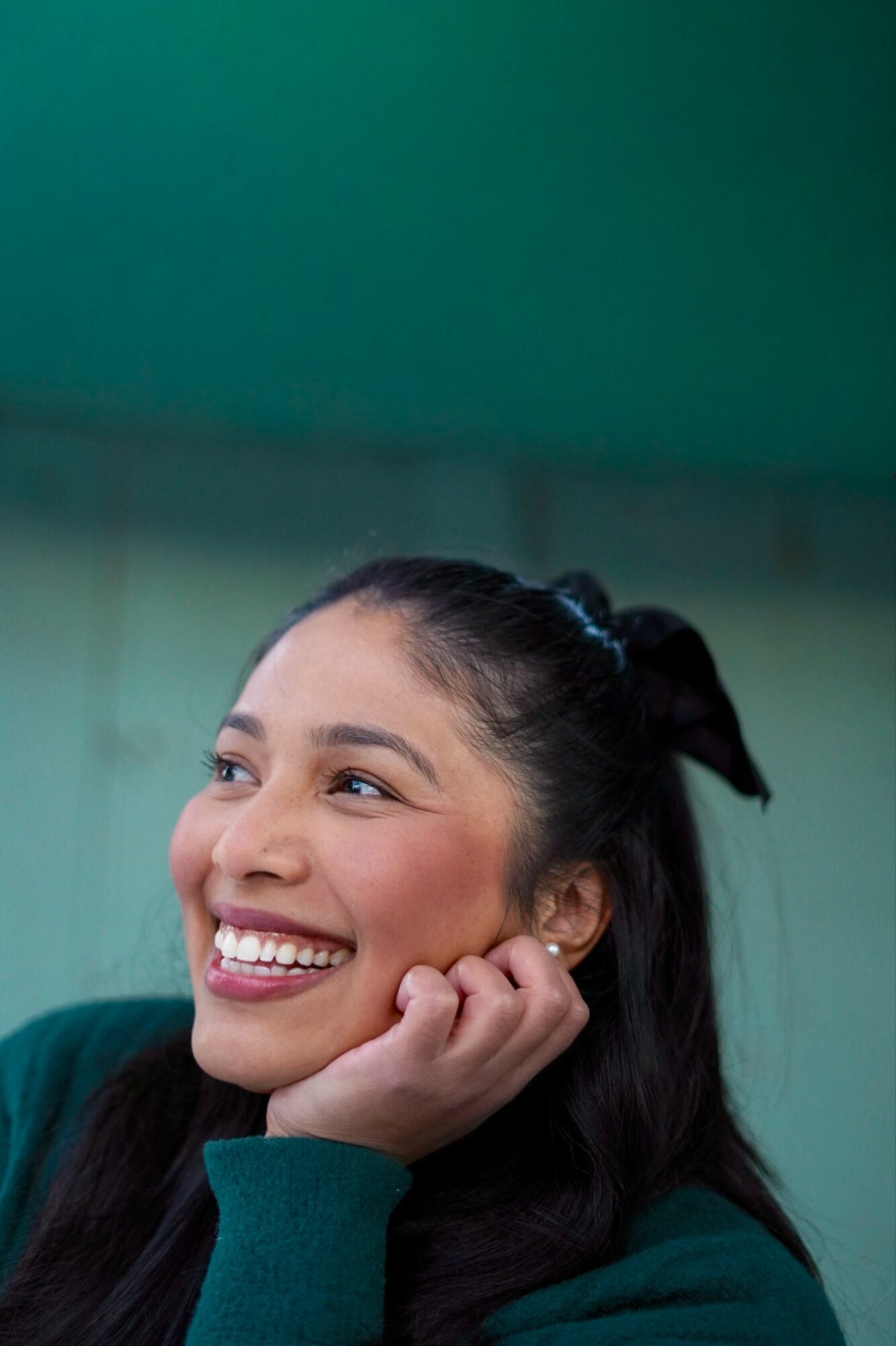 Smiling woman with dark hair tied back, resting her chin on her hand, against a green background.