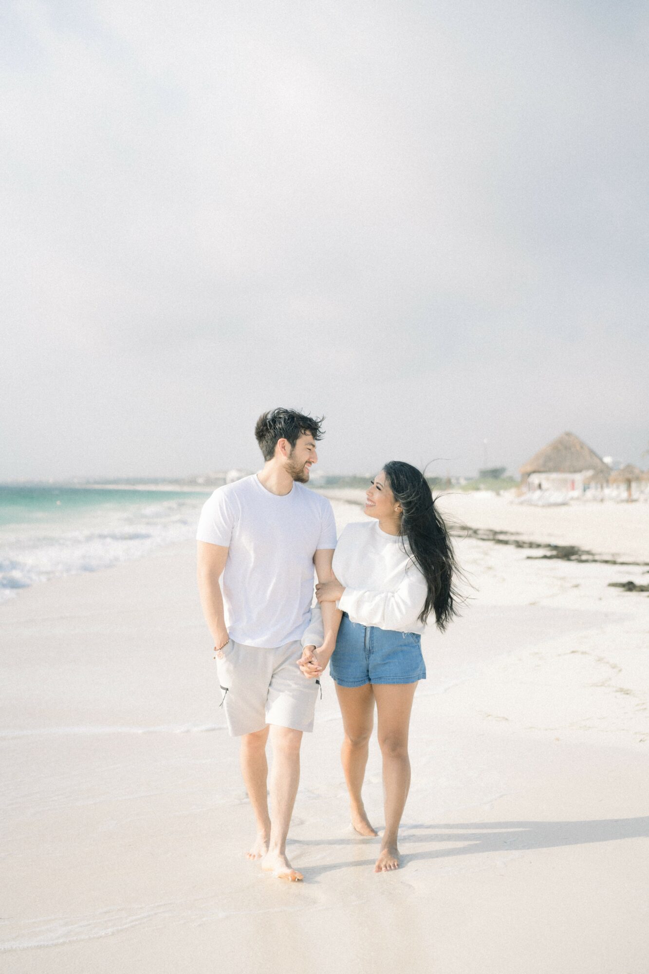A couple walking hand in hand on a beach, with the ocean and cloudy sky in the background.