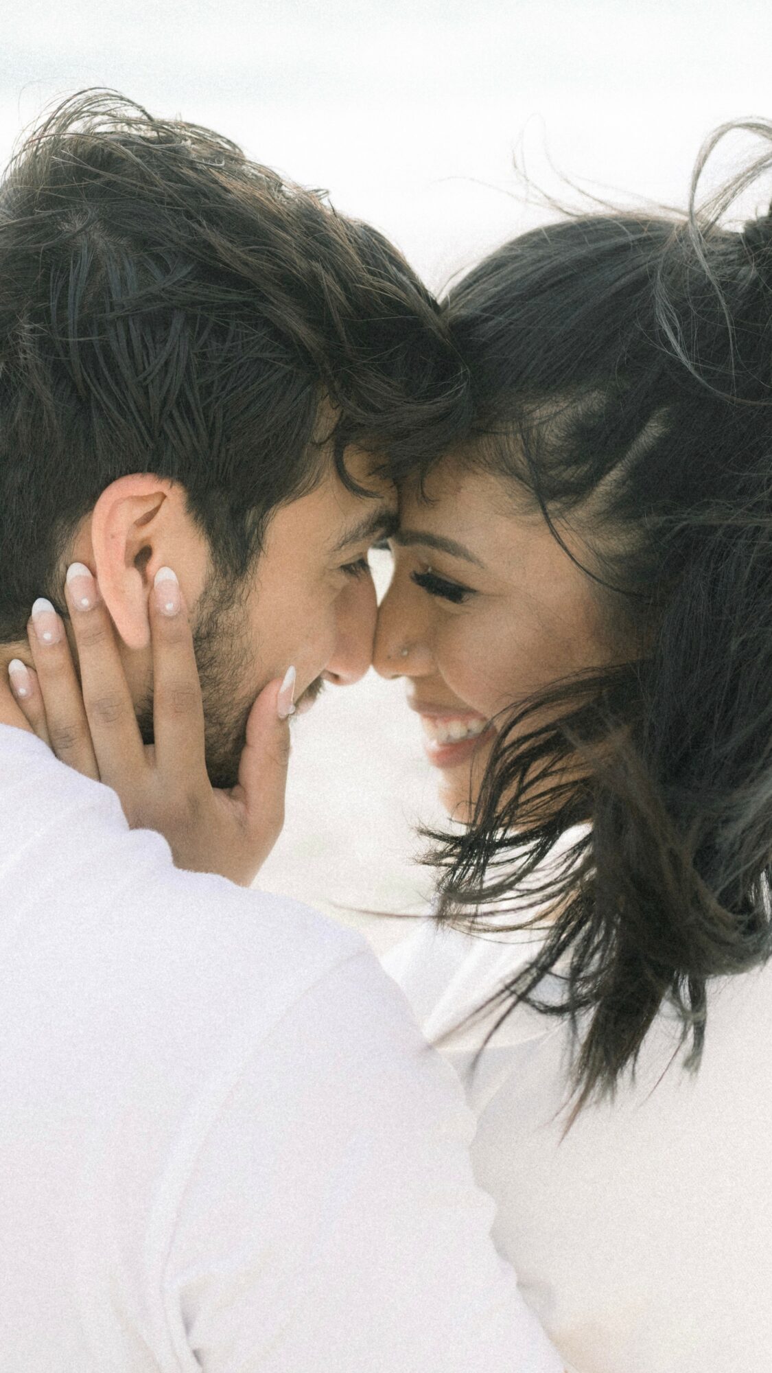Close-up of a man and woman touching foreheads and smiling, with their eyes closed, against a light background.