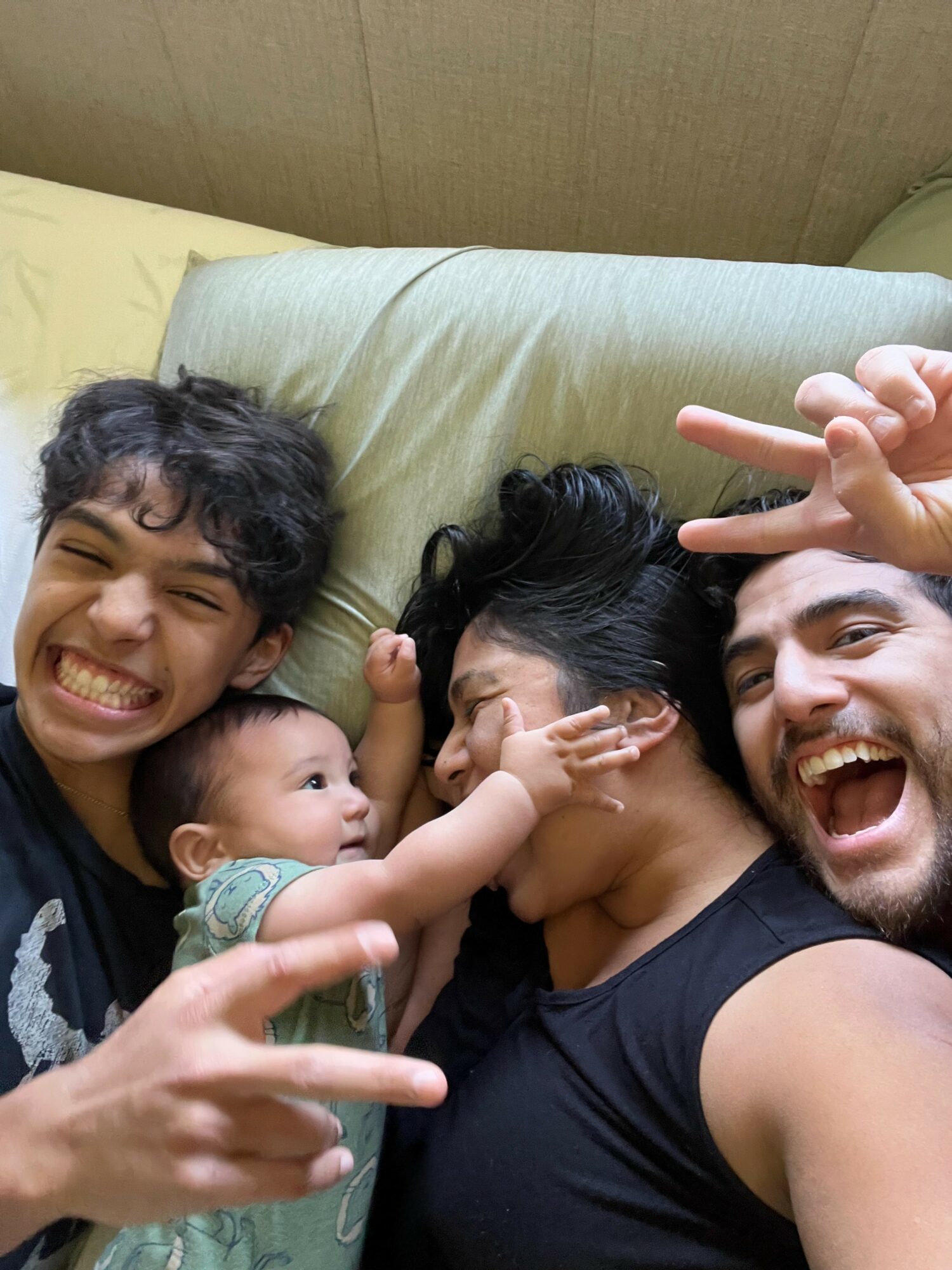 Four people lying on a bed, smiling and making peace signs, with a baby reaching towards a woman with black hair.