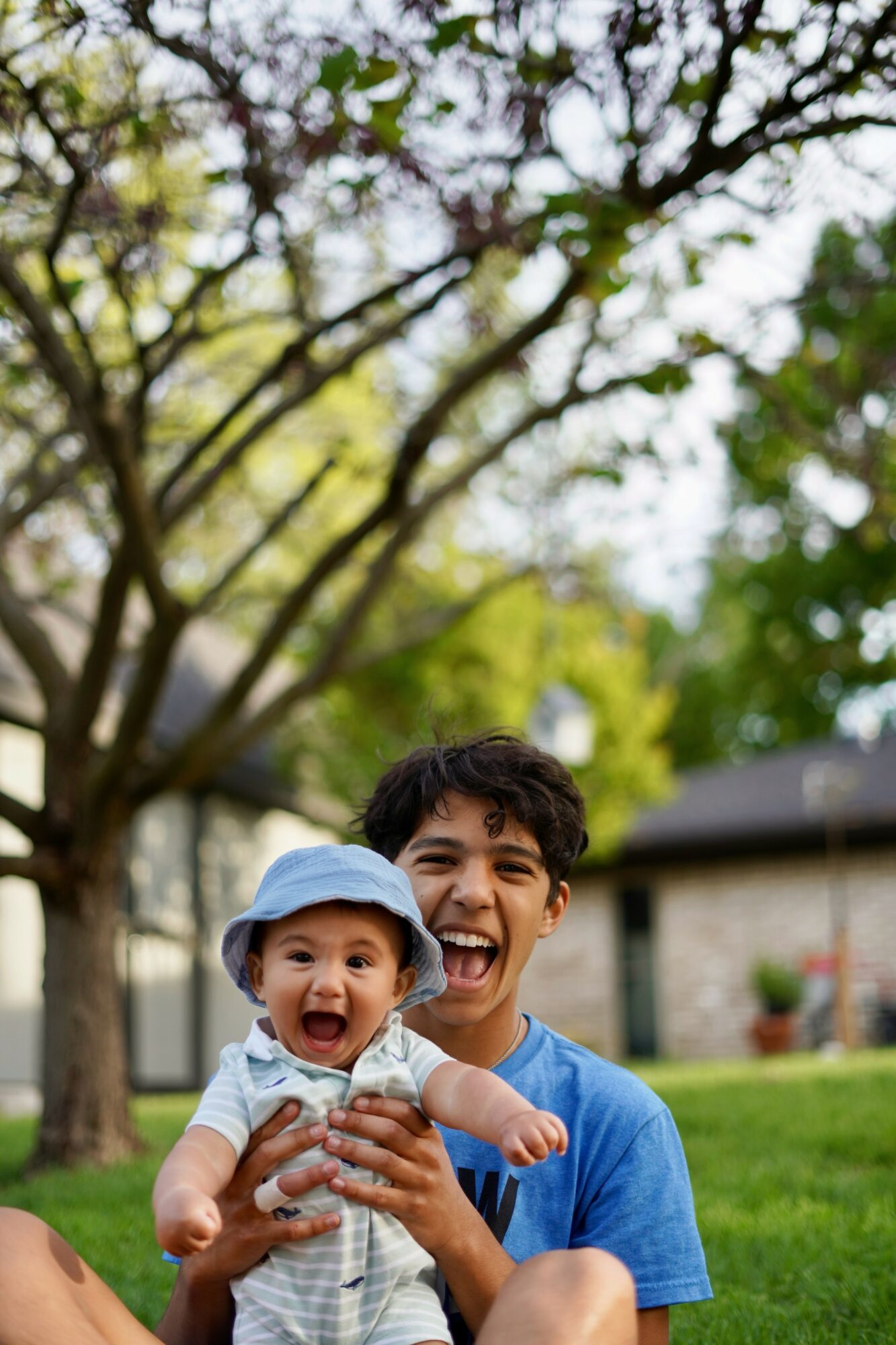 A young man holding a baby outdoors with trees and a building in the background.
