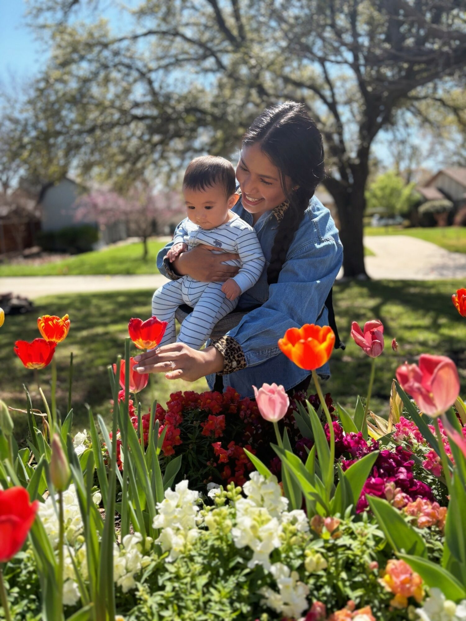 Woman holding a child in a garden with colorful flowers and trees in the background.