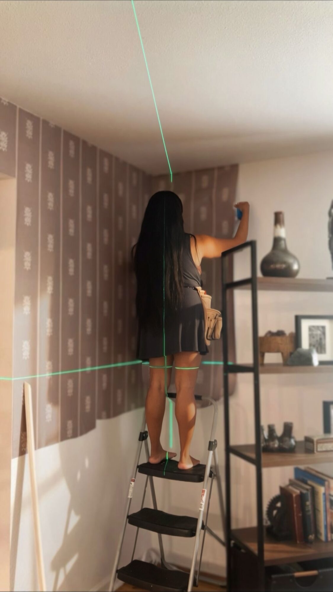 Person standing on a step ladder reaching for an object on a shelf, with patterned wallpaper and books nearby.