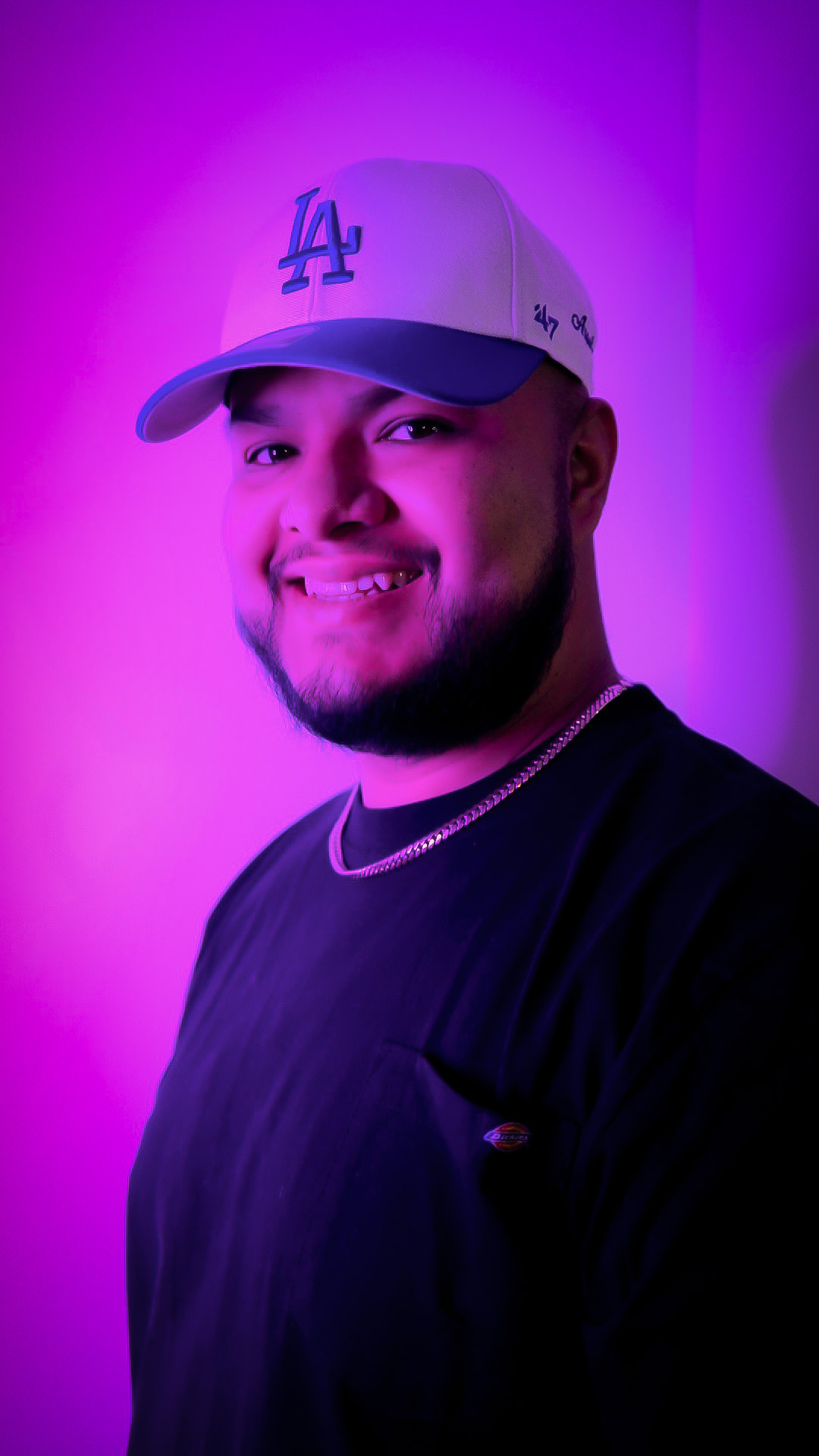 Young man smiling, wearing a white cap and black shirt, illuminated by purple lighting.