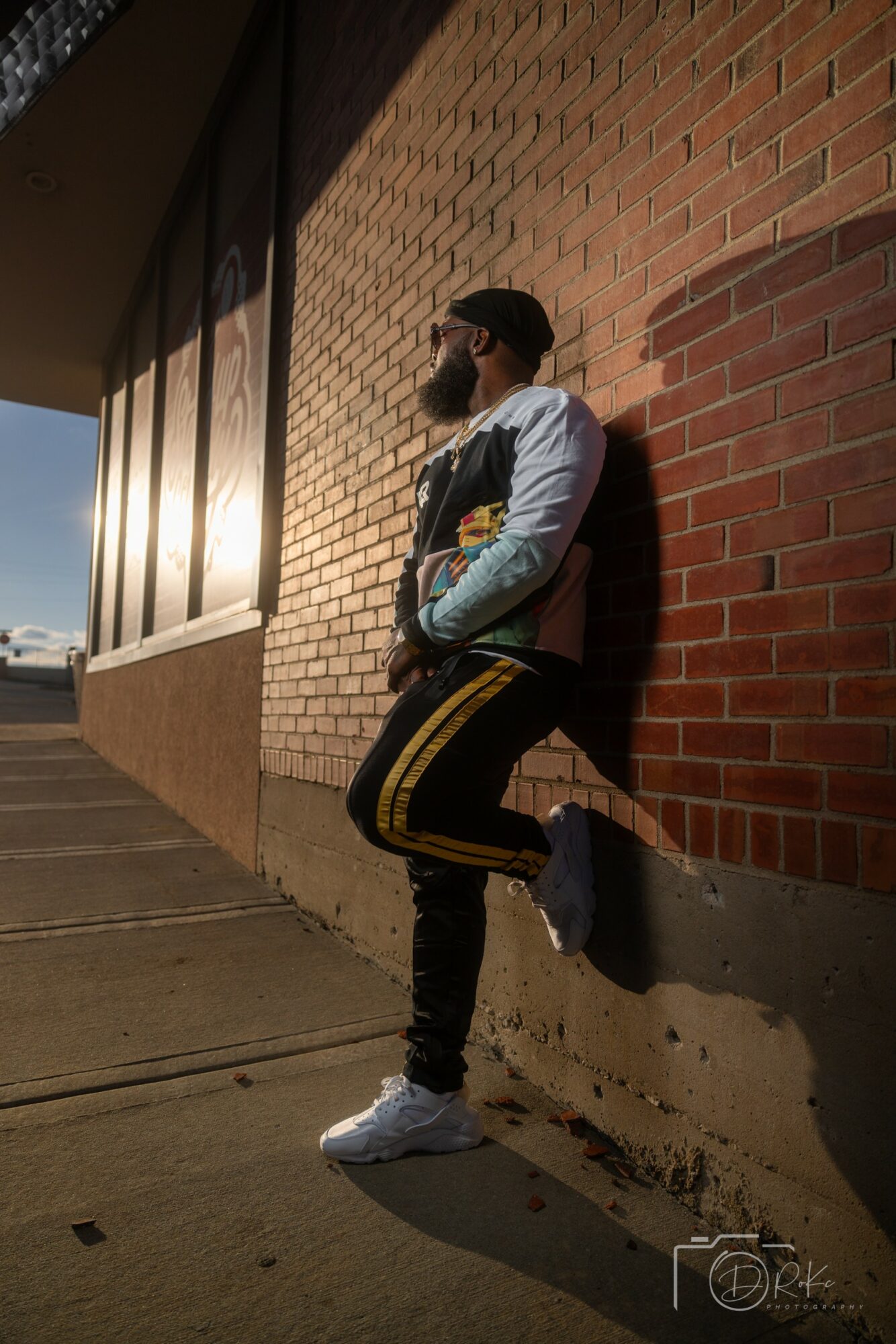 Person leaning against a brick wall during sunset, wearing a jacket, cap, and sneakers, with a cityscape in the background.