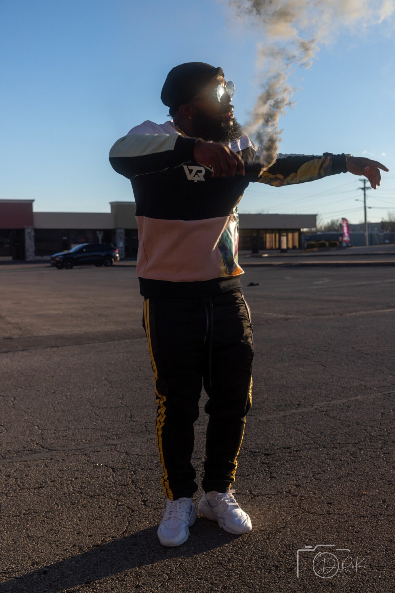 Person standing outdoors with arm extended, wearing a helmet, jacket, and pants, with a building and smoke in the background.