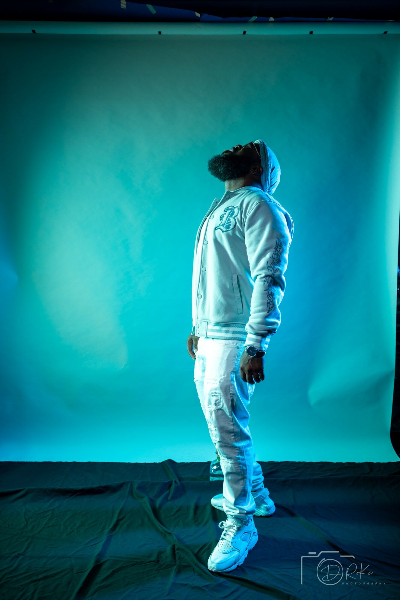 Person in hoodie and jeans standing sideways against a blue-green background, head slightly tilted upward.