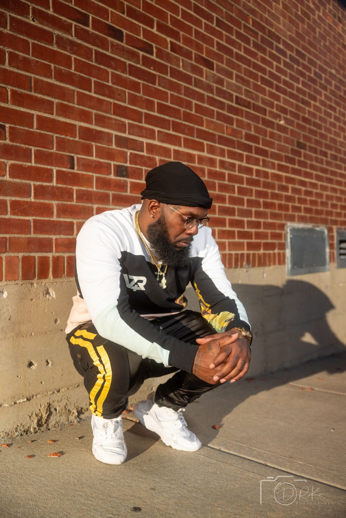 Person crouching on sidewalk against brick wall, wearing black beanie, glasses, white and black jacket, yellow-striped pants, and white shoes.