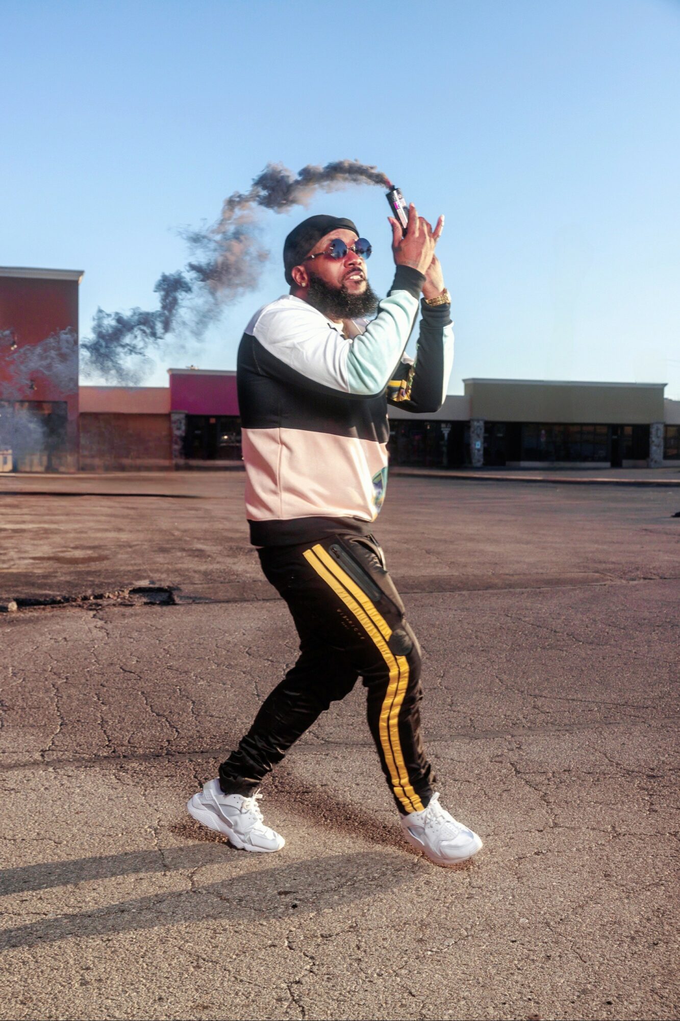 Person standing outdoors with hands together, wearing sunglasses, a jacket, and track pants, with a building and blue sky in background.