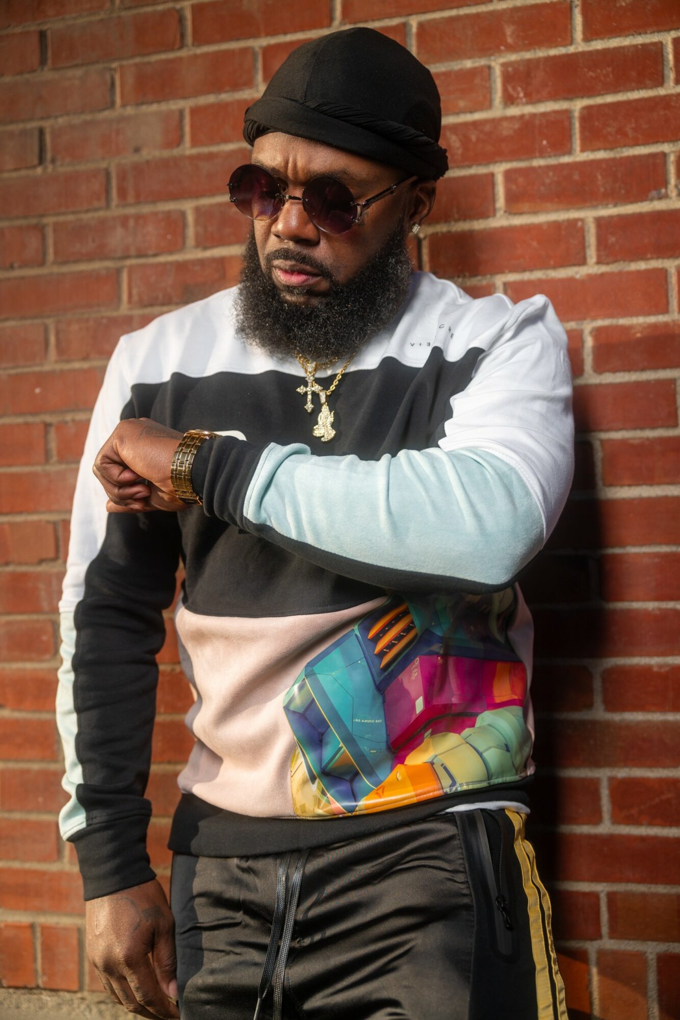 Man with sunglasses, black hat, and beard stands against a brick wall, wearing a colorful jacket and gold jewelry.
