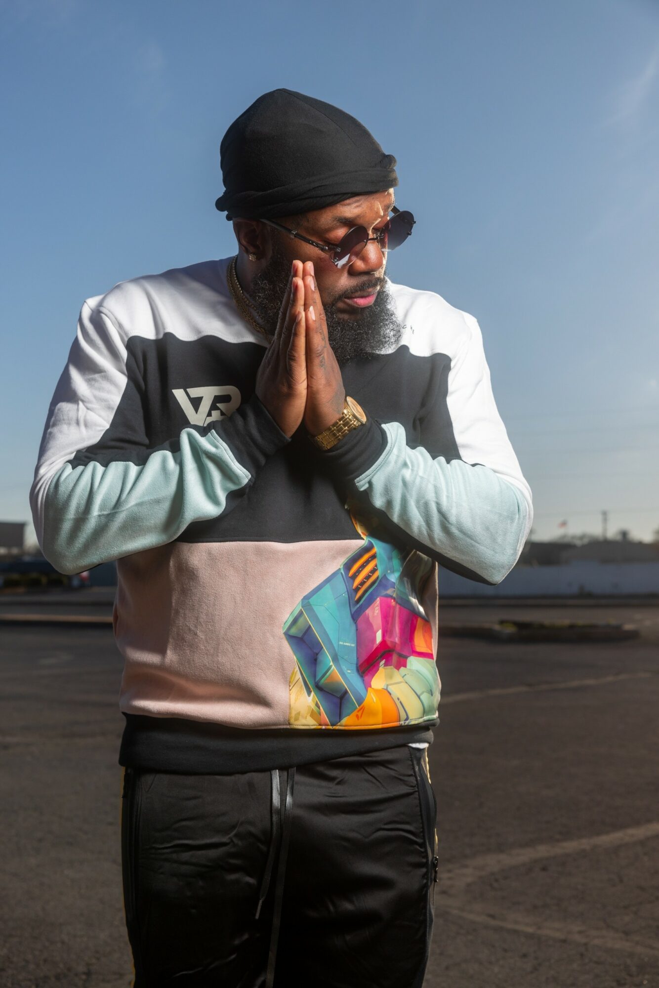 Man with glasses and a black head covering praying outdoors against a blue sky, wearing a jacket and colorful bag.