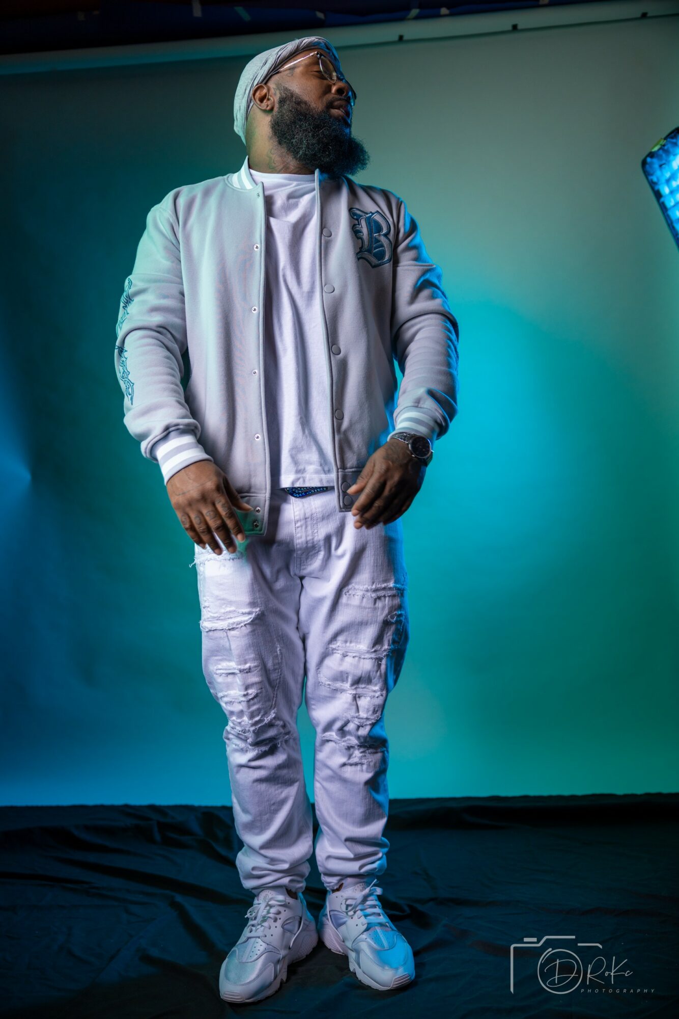 Man with a beard and glasses wearing a white hoodie and pants, standing against a blue-green background.