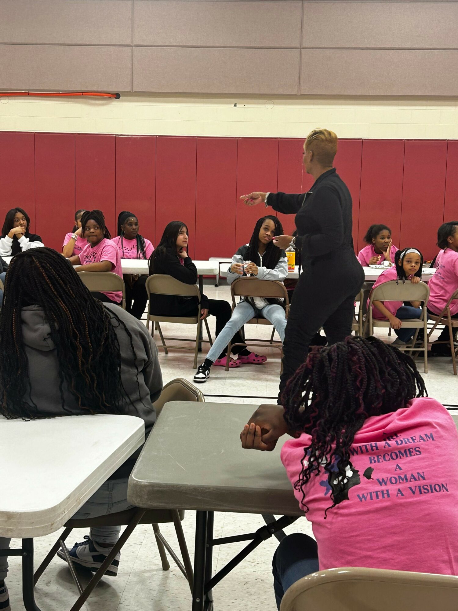 Group of people seated at tables in a room with red walls, some wearing pink shirts, others in black, engaging in a discussion.
