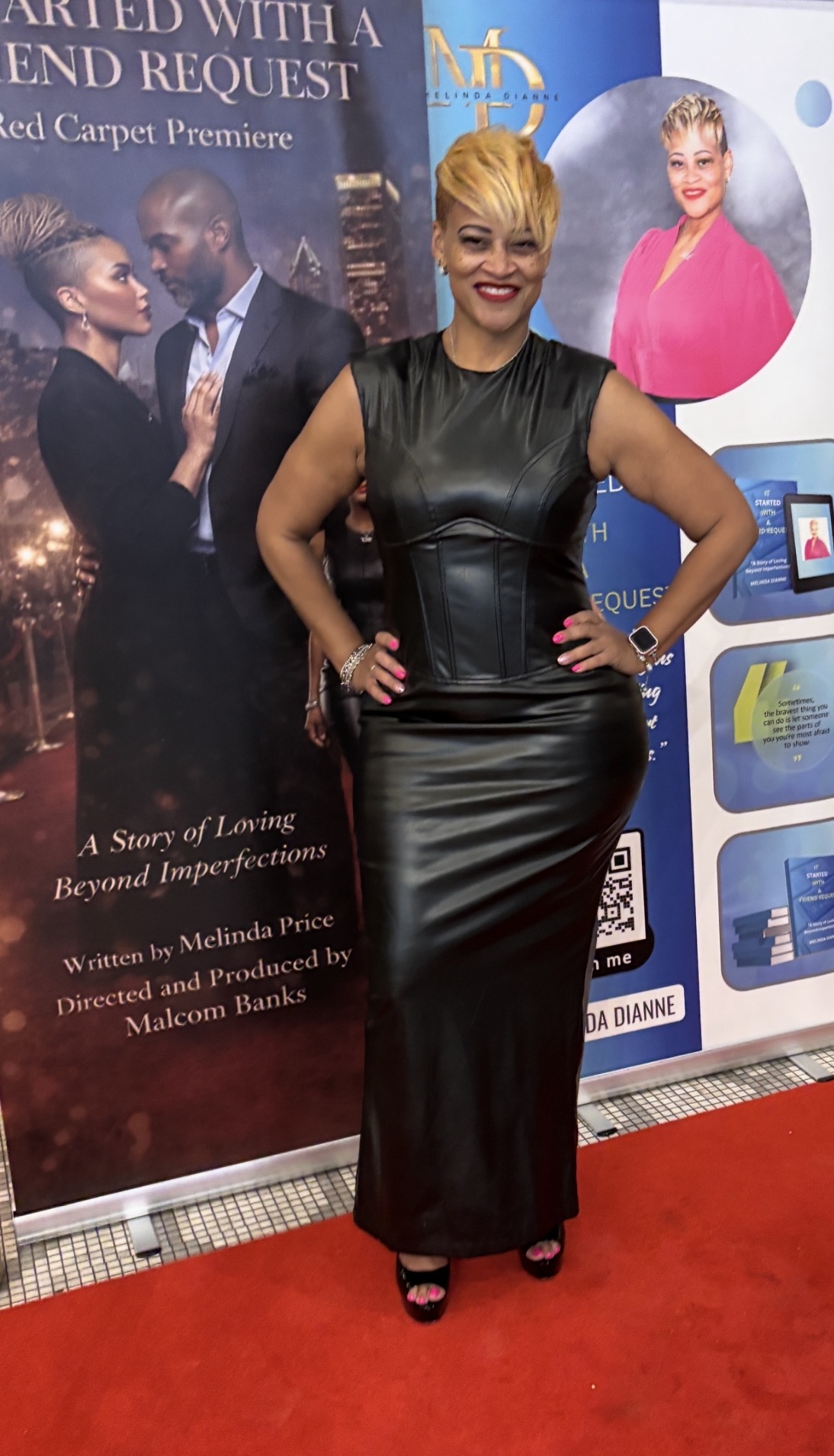 Woman with short blonde hair in a black leather dress standing on a red carpet, smiling, with promotional posters behind her.