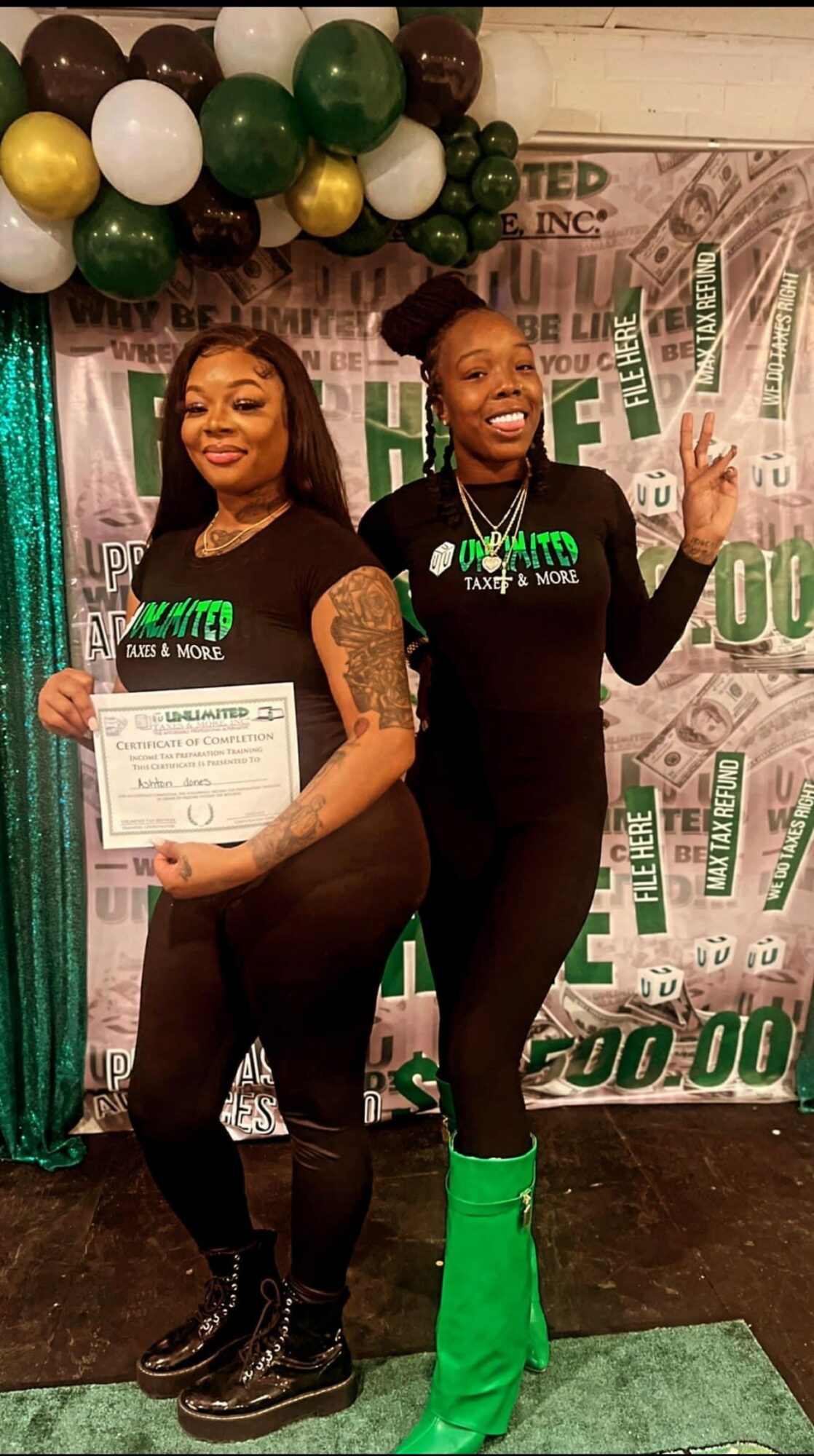 Two women standing in front of a decorated backdrop, one holding a certificate, both smiling and posing with peace signs.