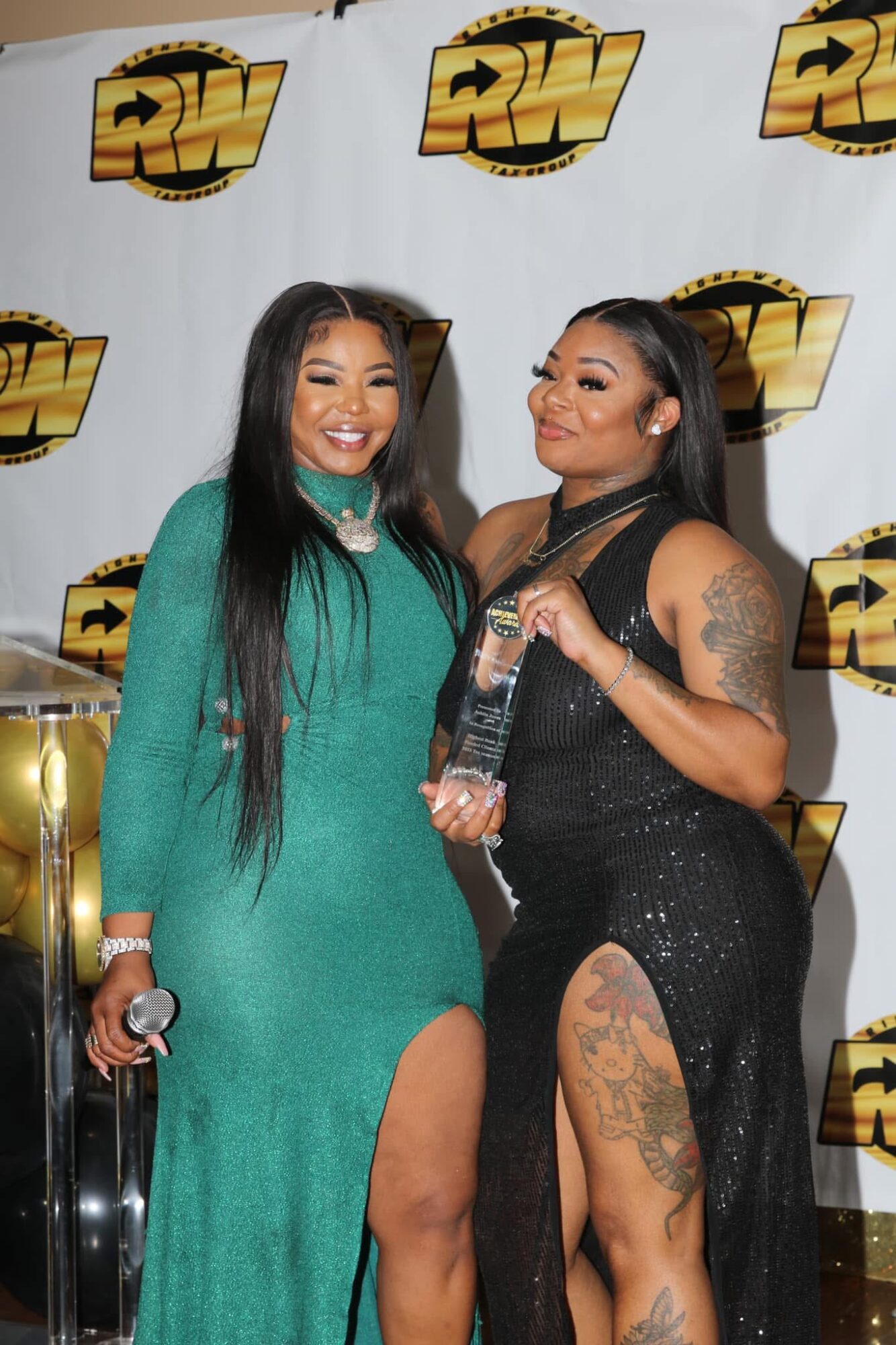 Two women smiling, one holding a trophy, standing in front of a backdrop with a logo, one wearing a green dress, the other in black.