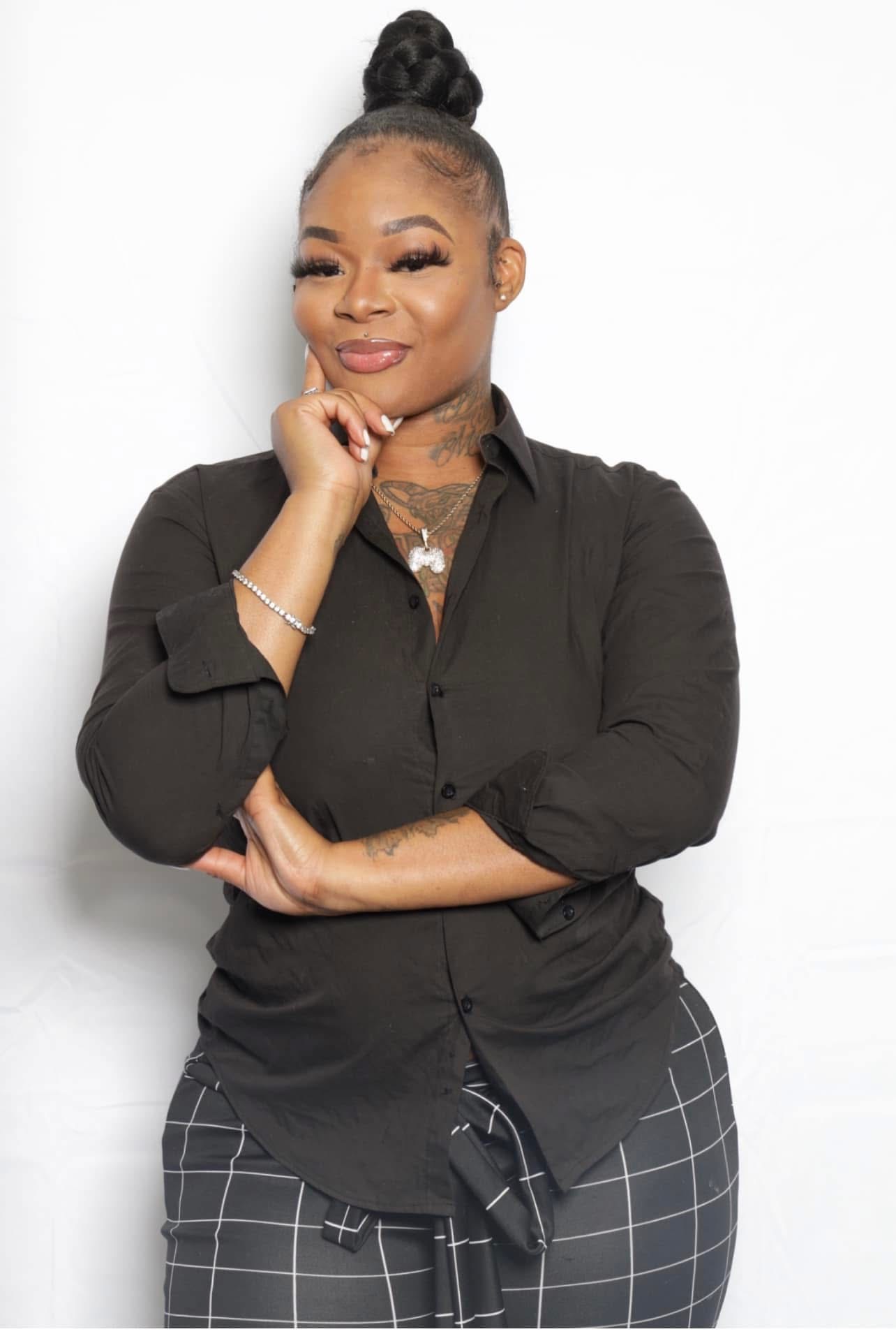 Woman with hair in a bun, smiling, wearing a black shirt and checkered pants, standing with arms crossed against a white background.