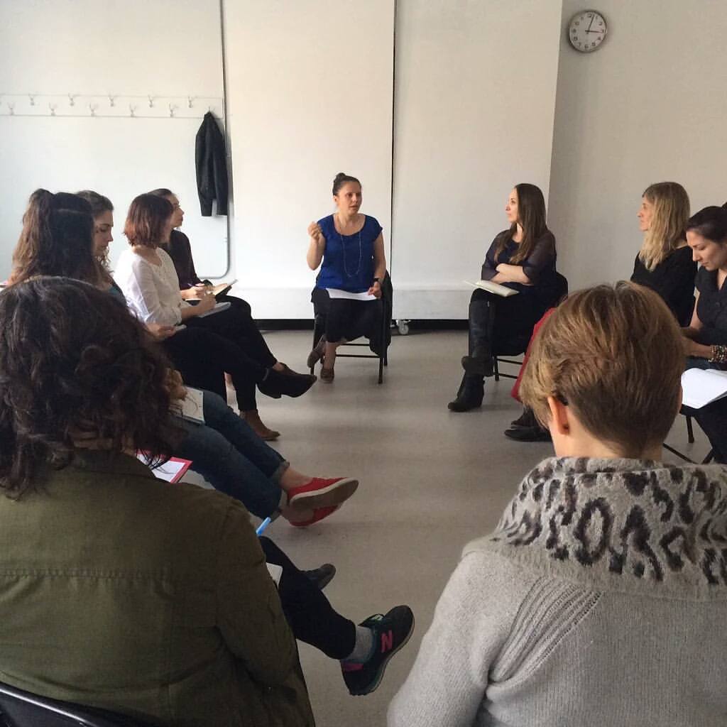 Group of people sitting in a circle in a room, engaging in a discussion or activity, with a clock on the wall.