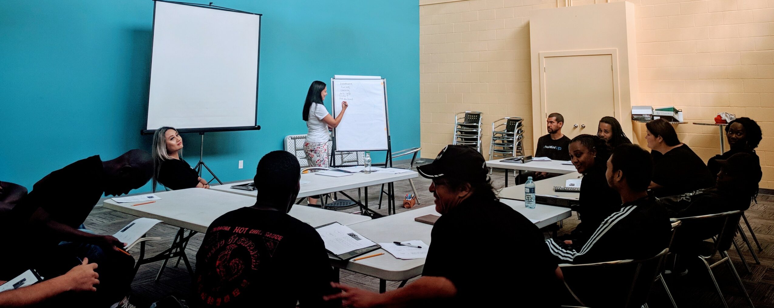 People attending a presentation in a conference room with blue and beige walls, seated around tables, a woman presenting at a whiteboard.