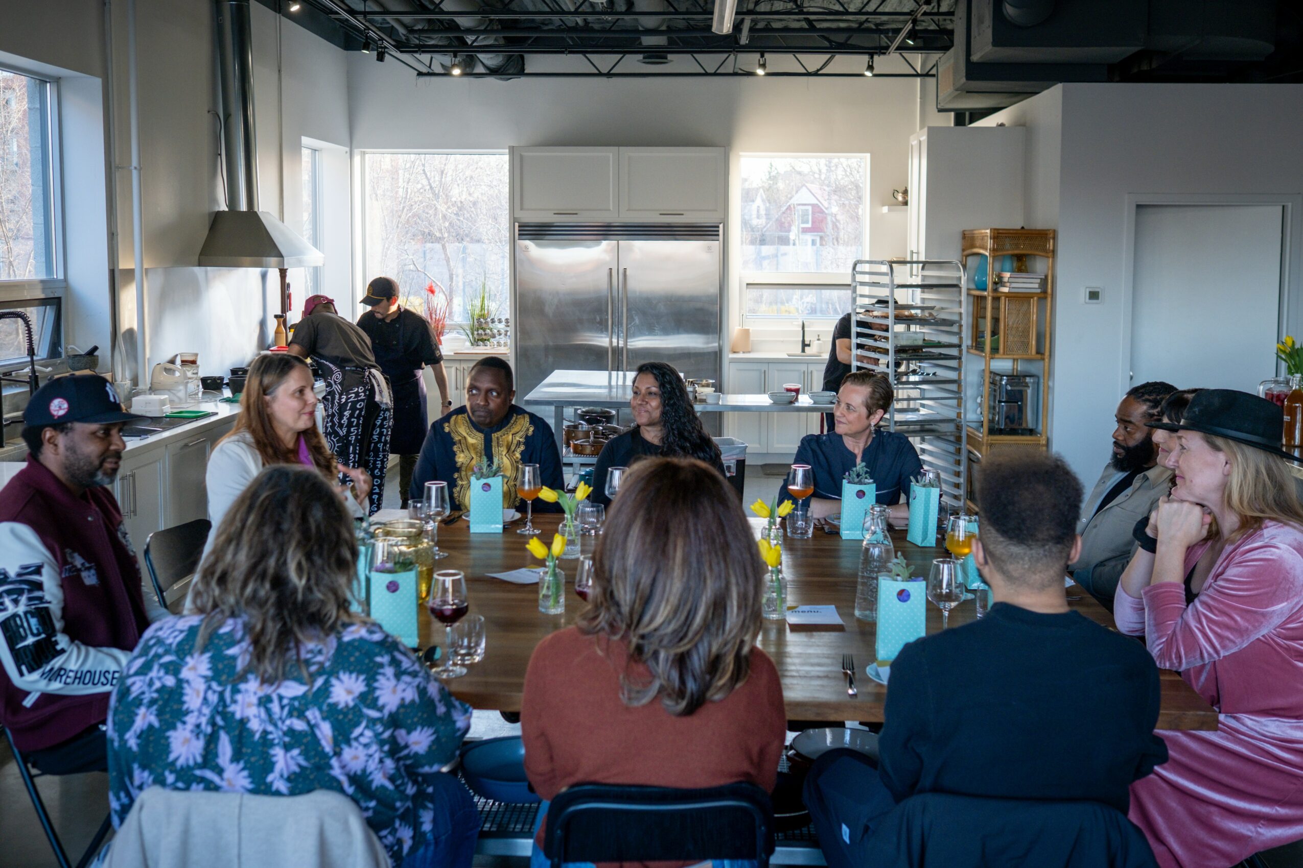 Group of people sitting around a table in a bright room, engaging in conversation, with drinks and snacks present.