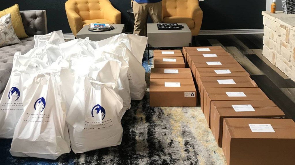 Multiple white bags and stacked brown boxes on a patterned rug in a living room with chairs and a table.
