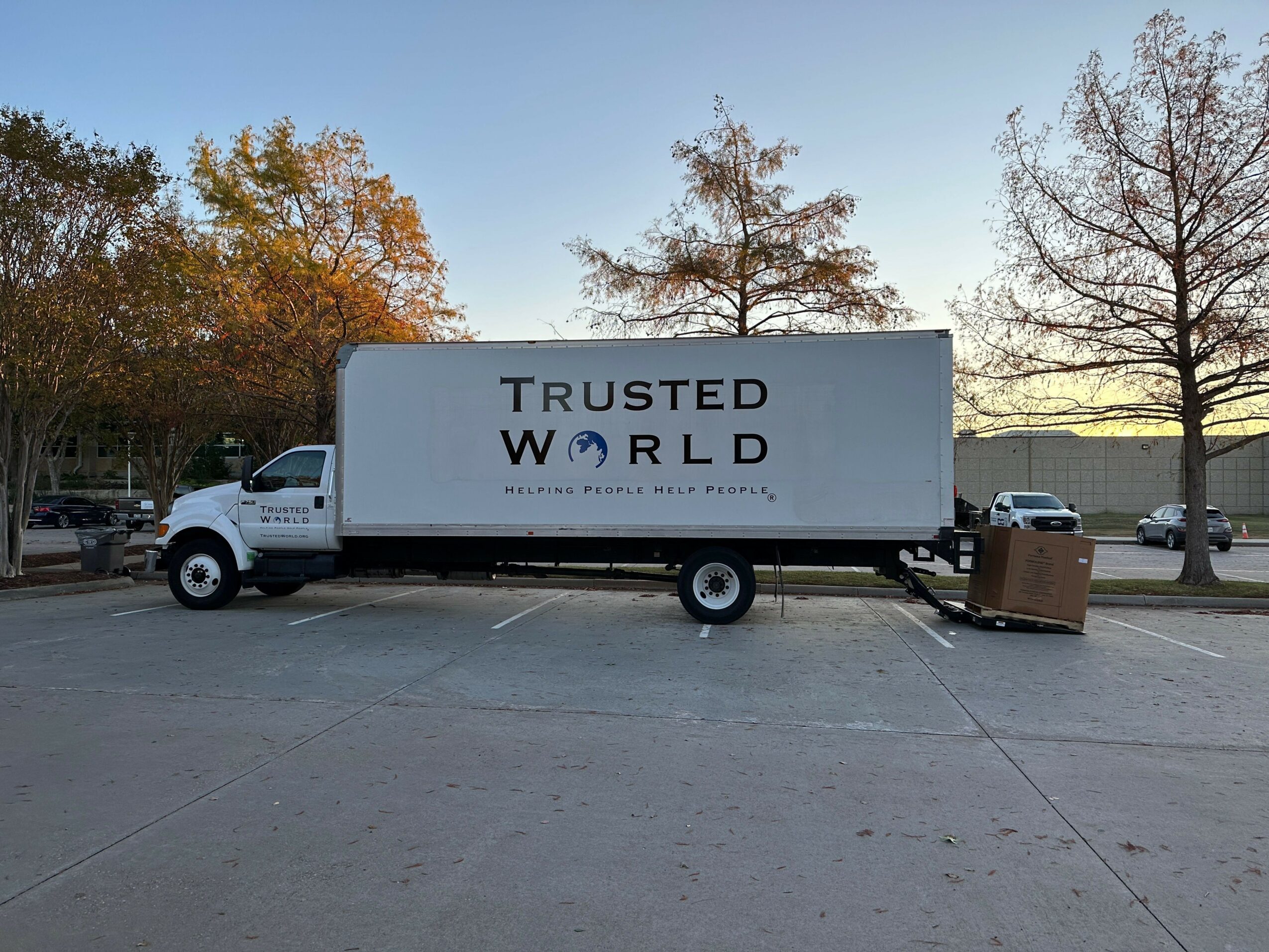 A truck with a sign reading 'TRUSTED WORLD' is parked on a road with trees and a clear sky in the background.