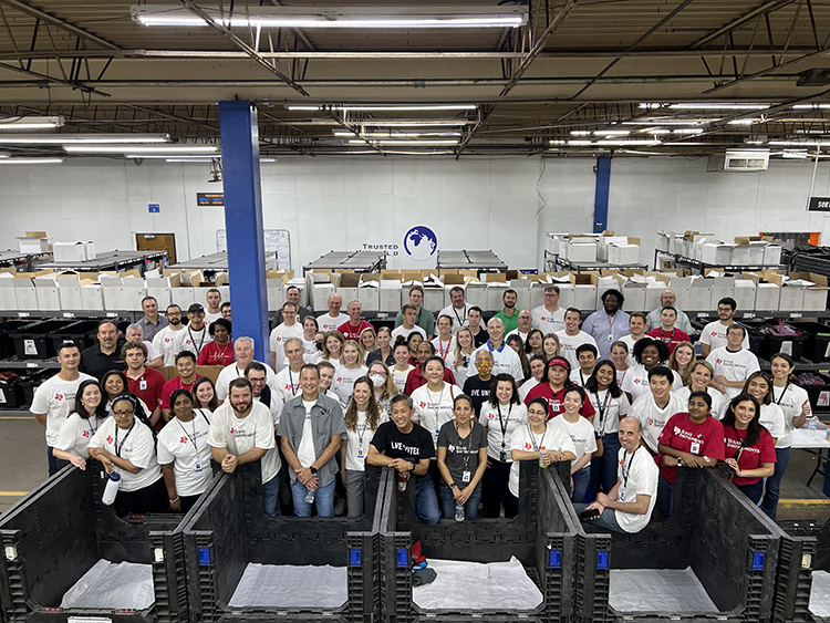 Group of people in a warehouse with shelves and equipment, some wearing uniforms, standing and sitting in front of workstations.