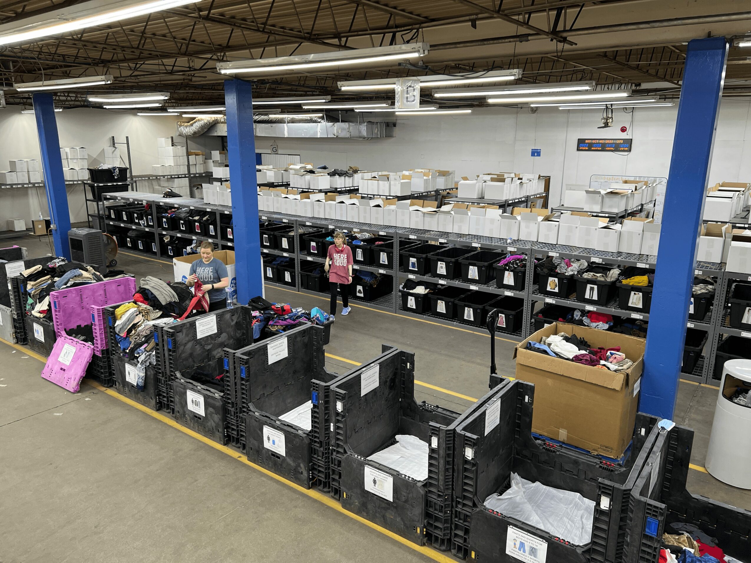 Warehouse with shelves, bins, and workers organizing items, blue support columns, and industrial ceiling, viewed from the front.