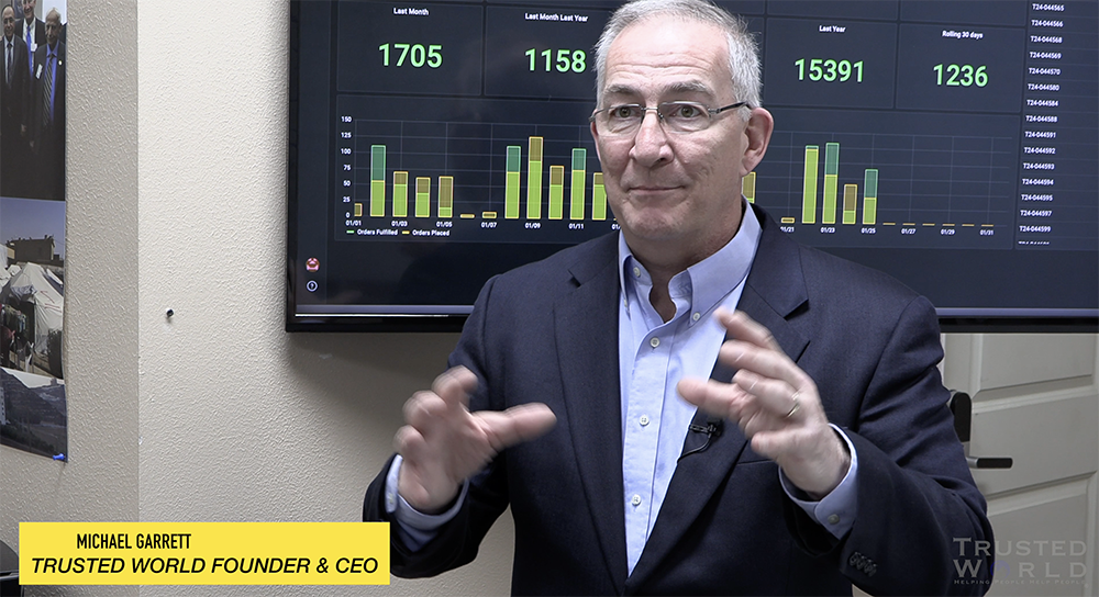 Man in suit speaking in front of a screen with graphs, gesturing with hands, in an office setting.
