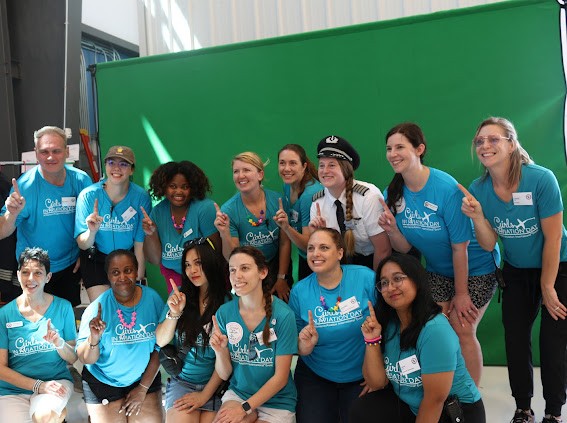 Group of women and girls wearing matching blue shirts, posing in front of a green screen, smiling and raising fists.