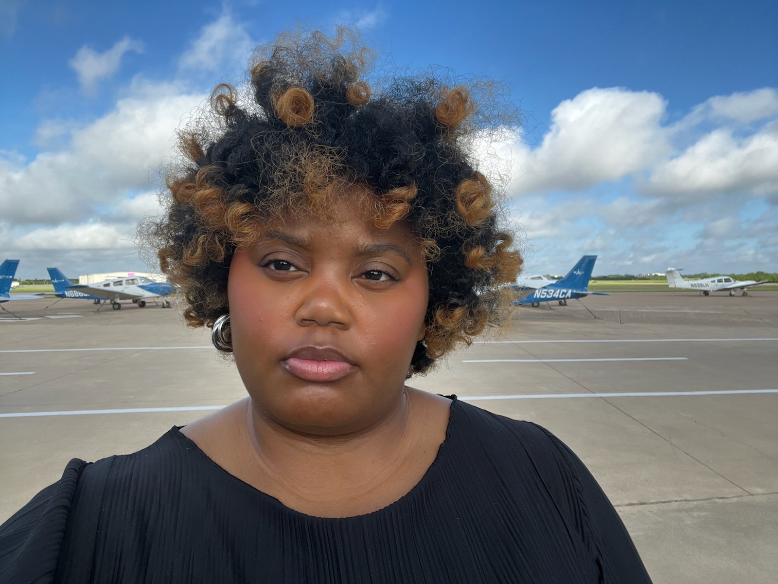 Young woman with curly hair standing on an airport tarmac with planes in the background under a partly cloudy sky.