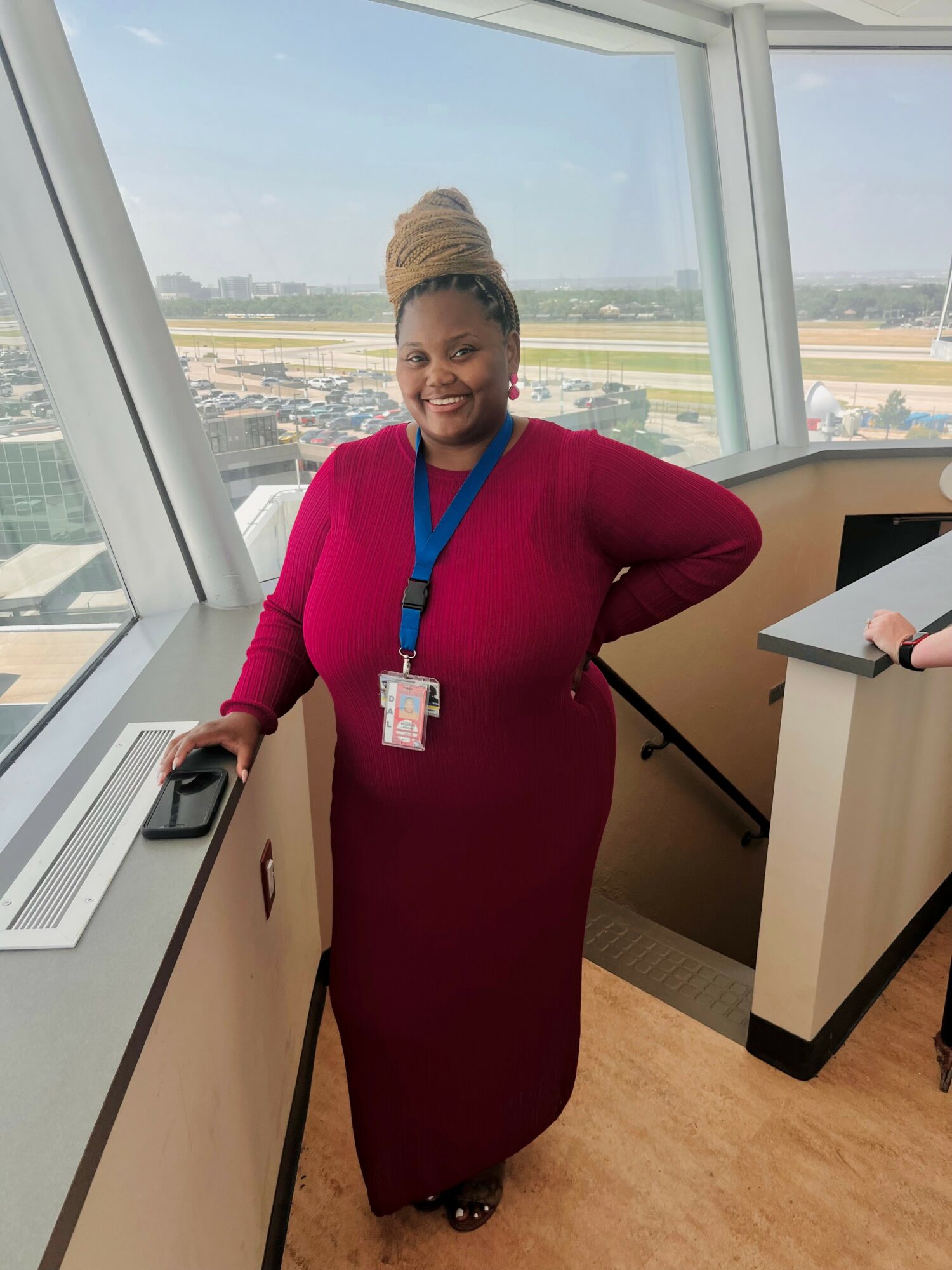 Woman in a long red dress with a beige headwrap, smiling, standing inside an airport terminal near large windows, holding a phone.