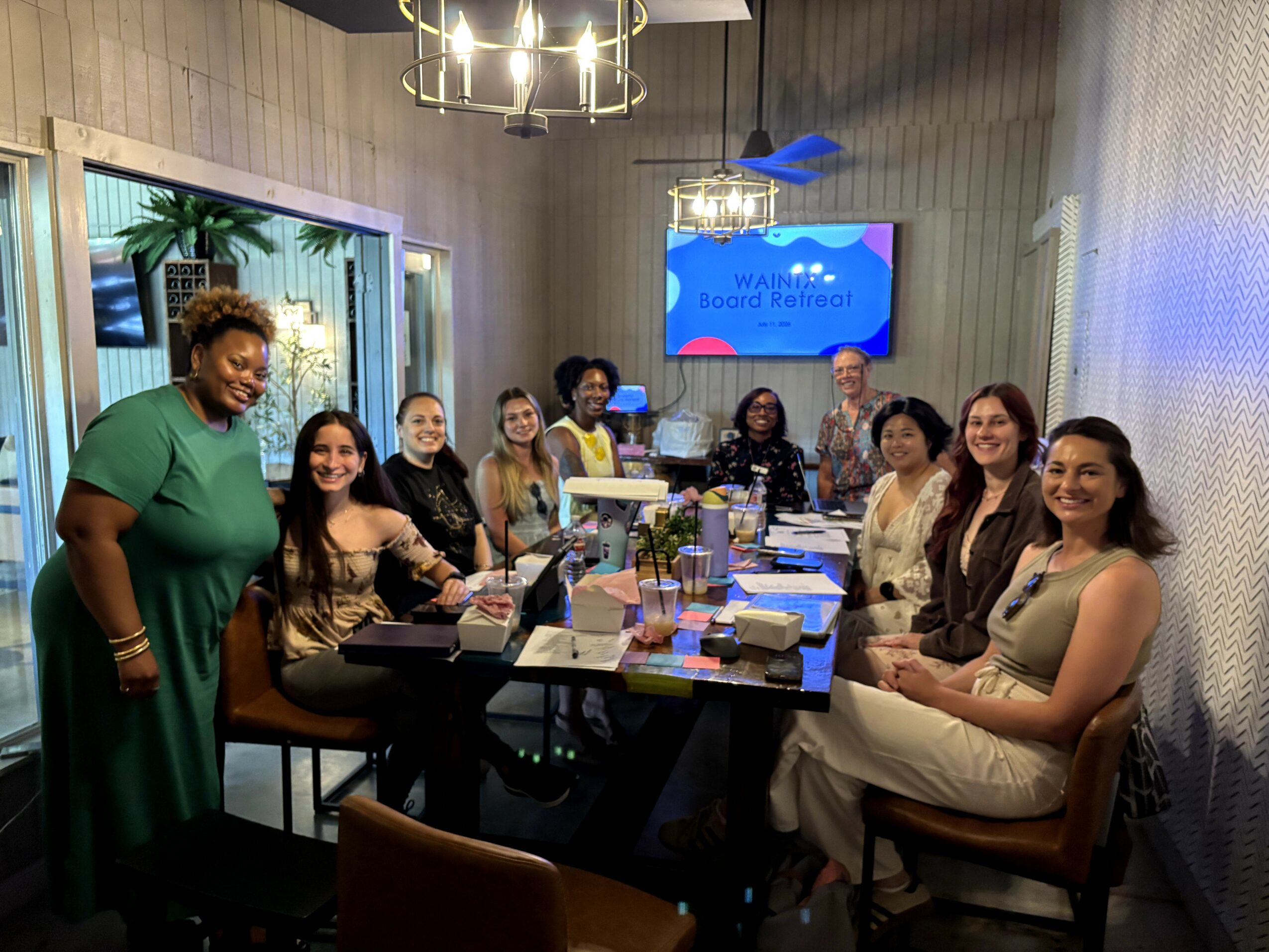Group of ten women sitting around a conference table in a modern room, smiling at the camera, with a screen displaying a presentation in the background.