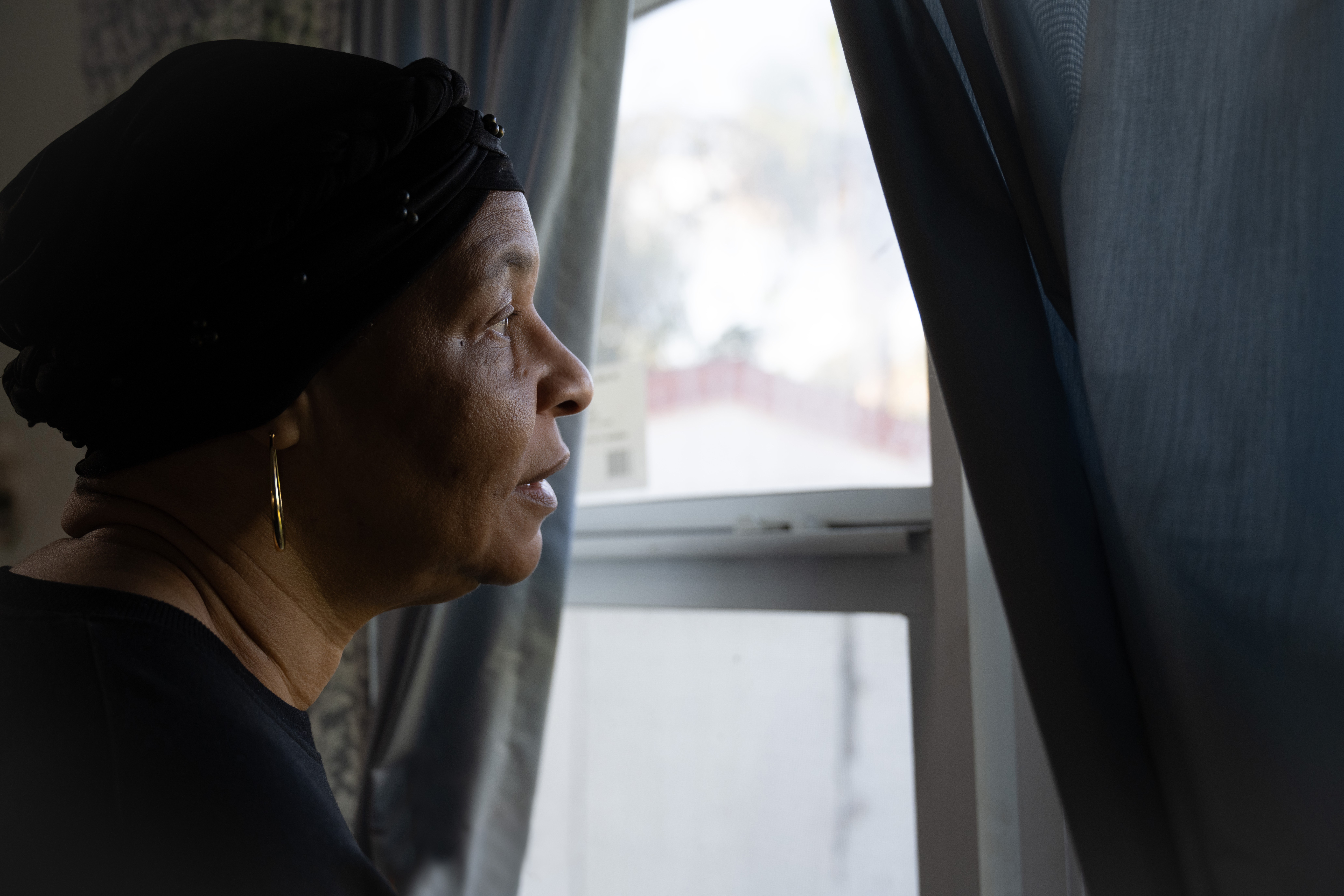Woman looking out of a window with curtains partially drawn, profile view, indoor setting.