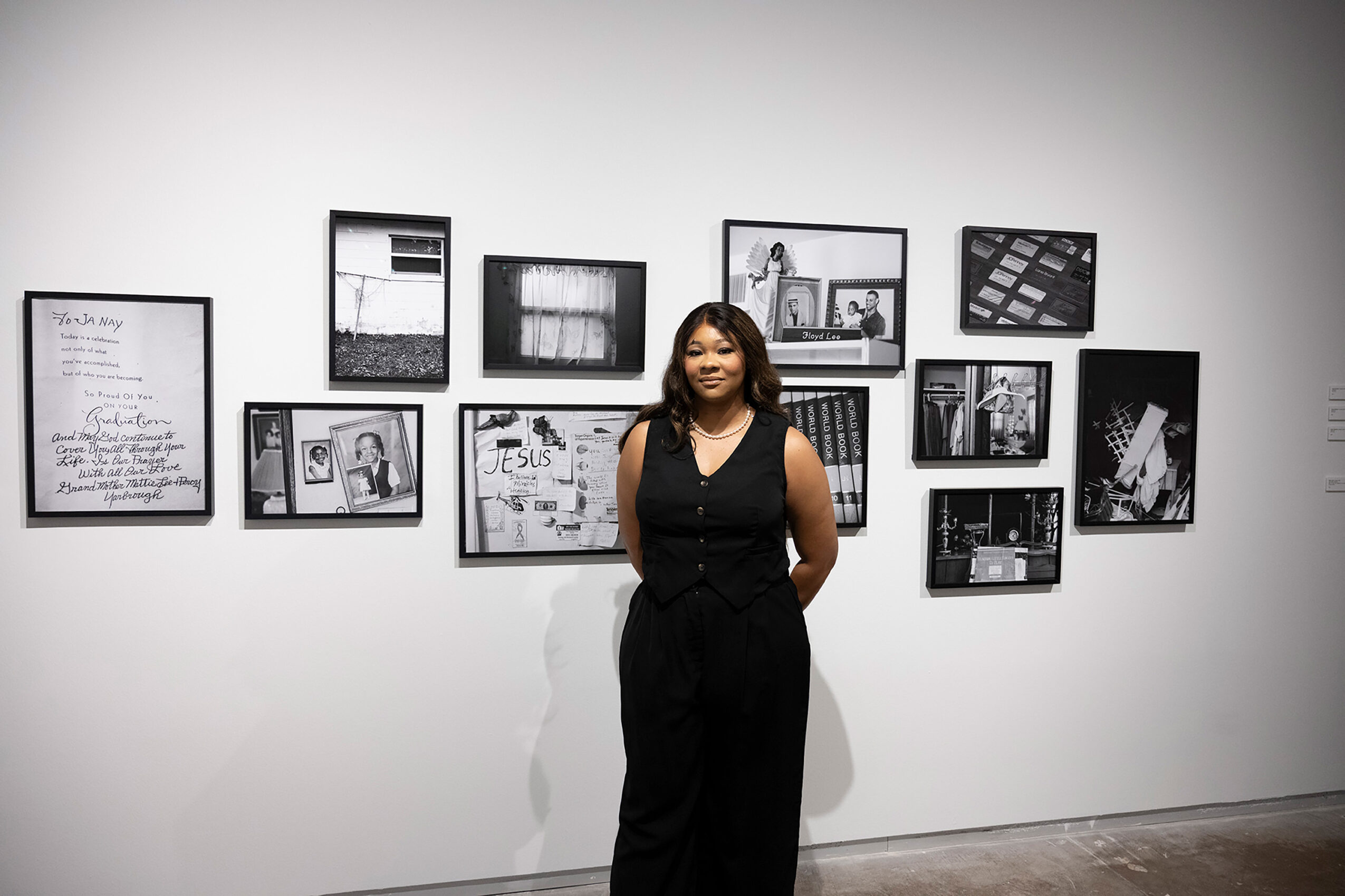 Woman standing in front of a wall with framed black-and-white photographs and artwork, smiling, with hands behind her back.