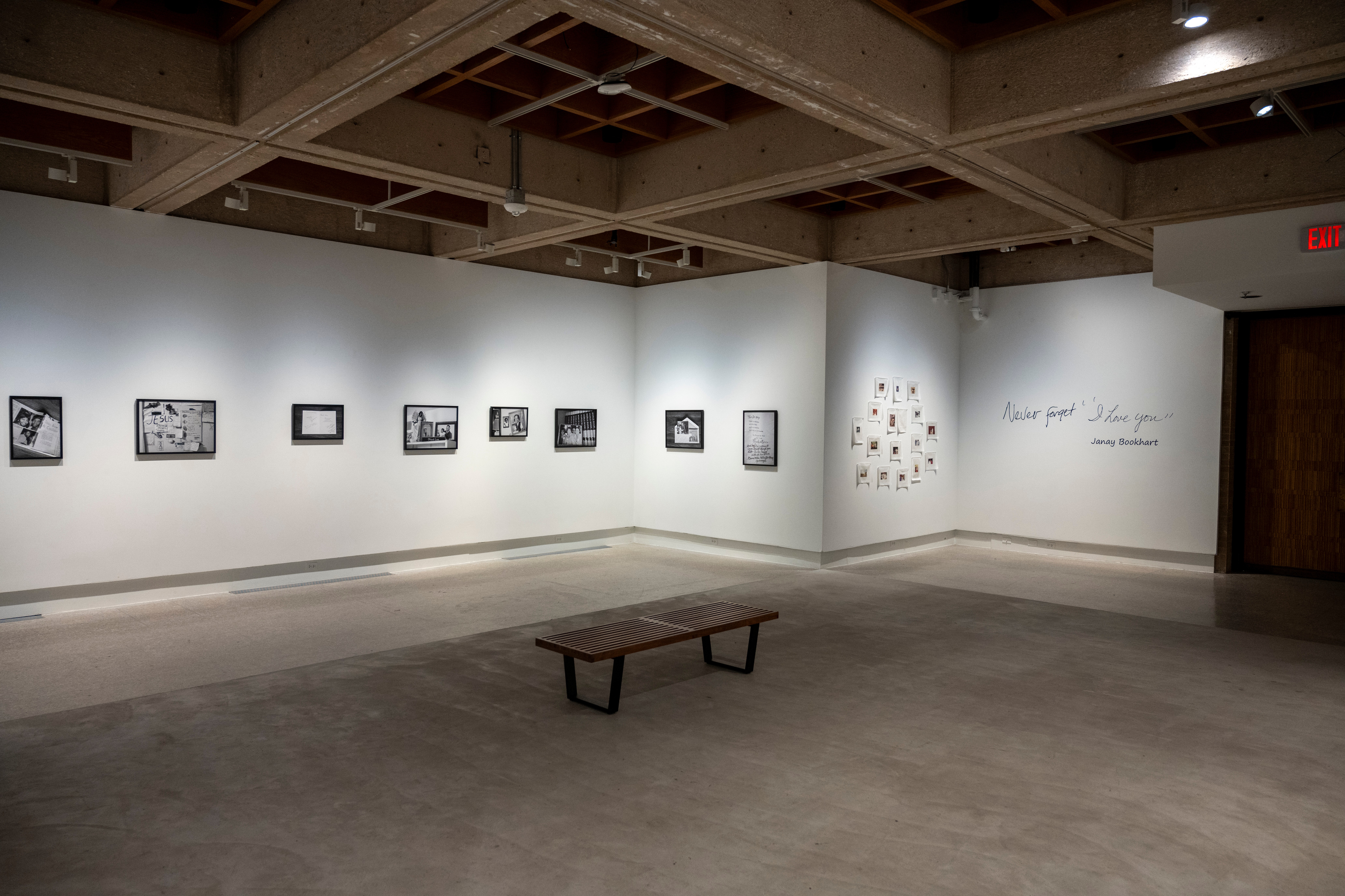 Gallery with framed artwork on white walls, a bench in the center, and a wall with text and small objects, empty space.