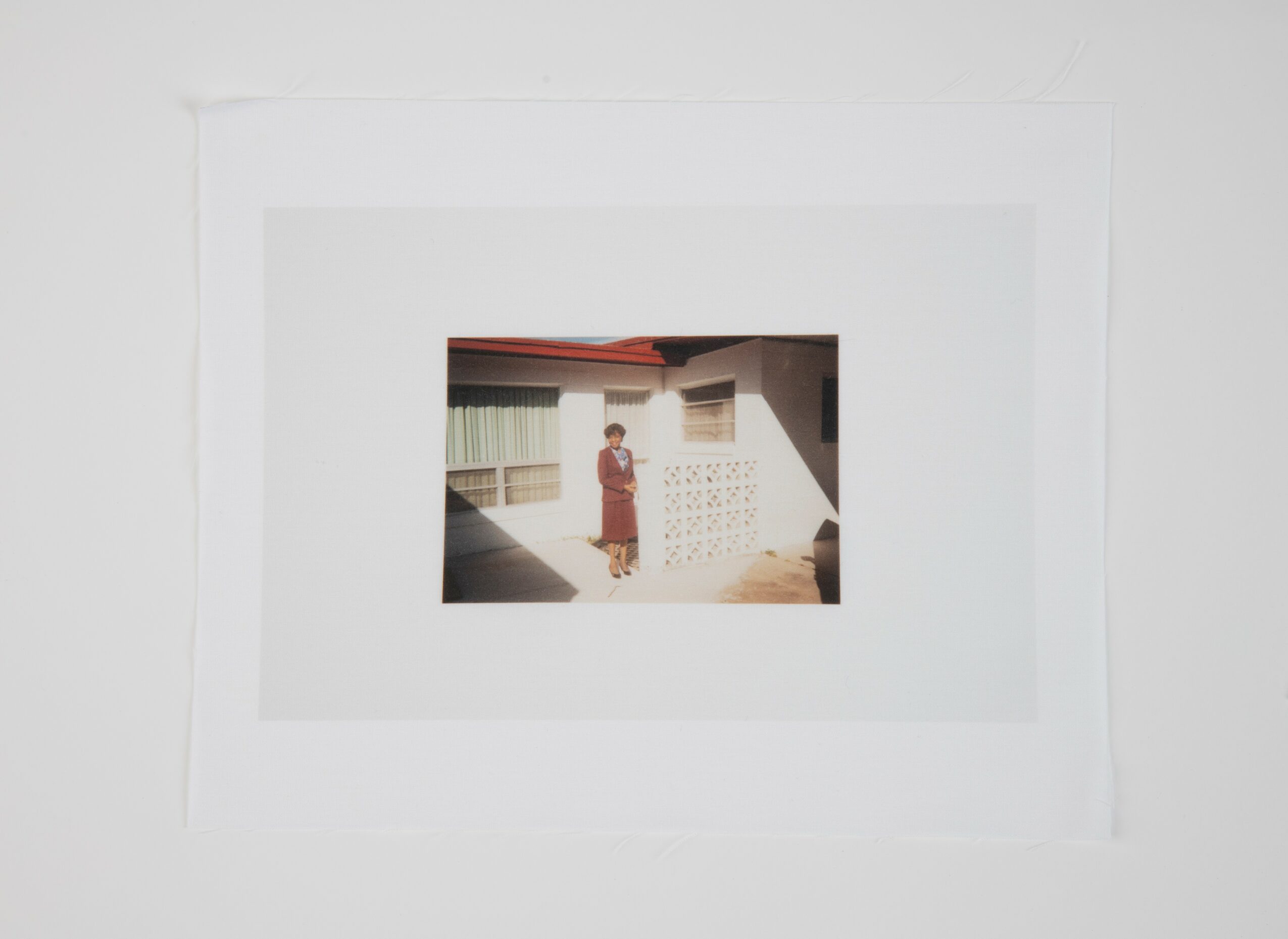 Person standing outside a building with a window and a wall, sunlight casting shadows, in a residential area.