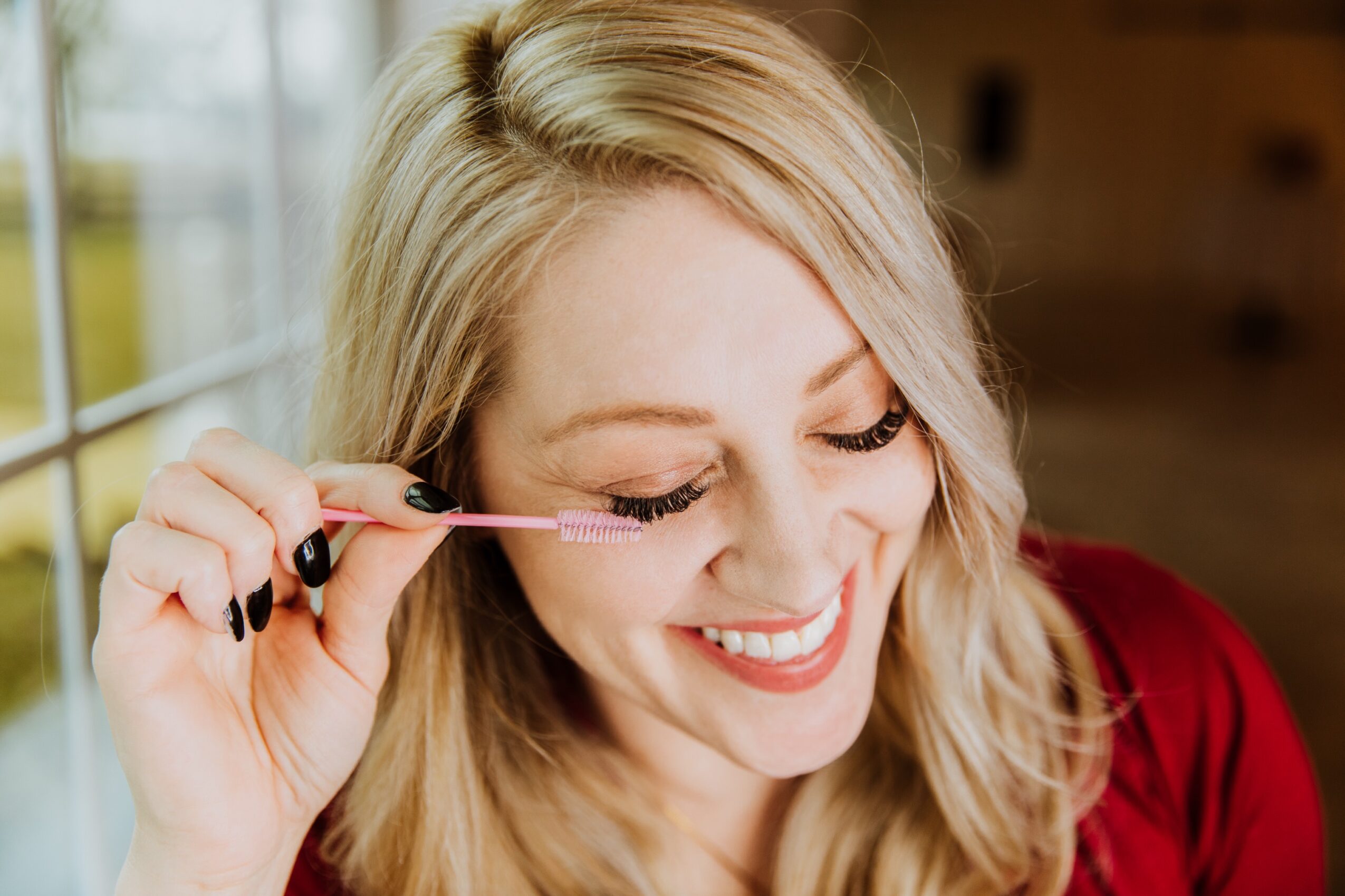 Woman applying mascara, smiling, with blonde hair and red top, near a window with natural light.